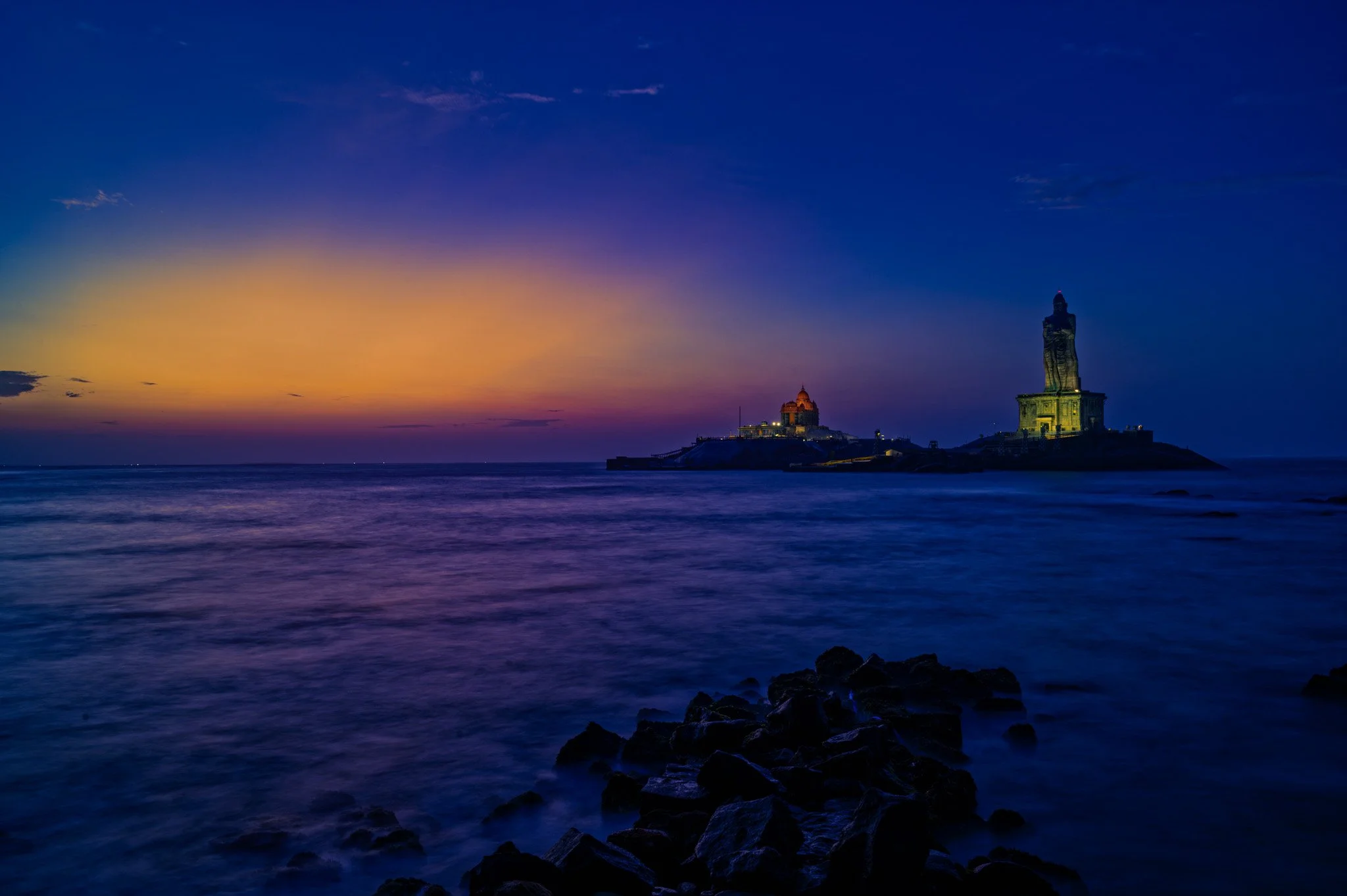 A long-exposure photograph of the Thiruvalluvar Statue at dawn in Kanyakumari, where the ocean waves blur into a misty white dreamscape with stunning exhibition detail.