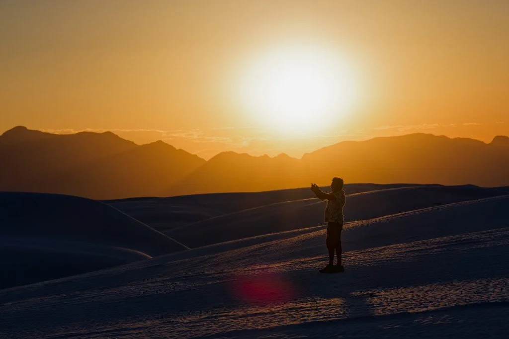 A dramatic silhouette of a lone traveler standing on a dark dune ridge, holding a camera against a massive, glowing golden sun as it sets behind distant mountain peaks.