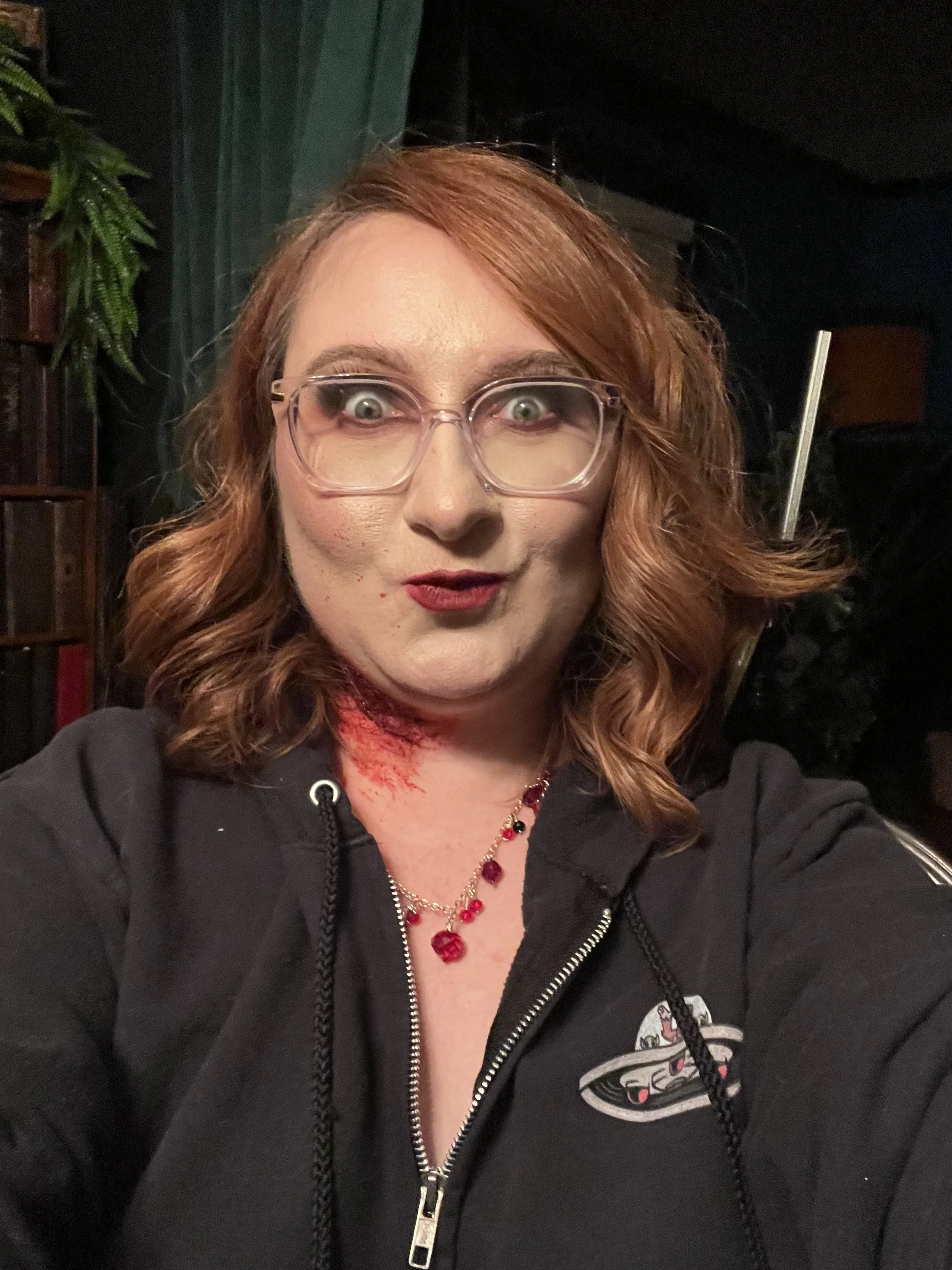 Woman with red hair and crimson makeup making a silly face at the camera with fake blood smeared on her neck