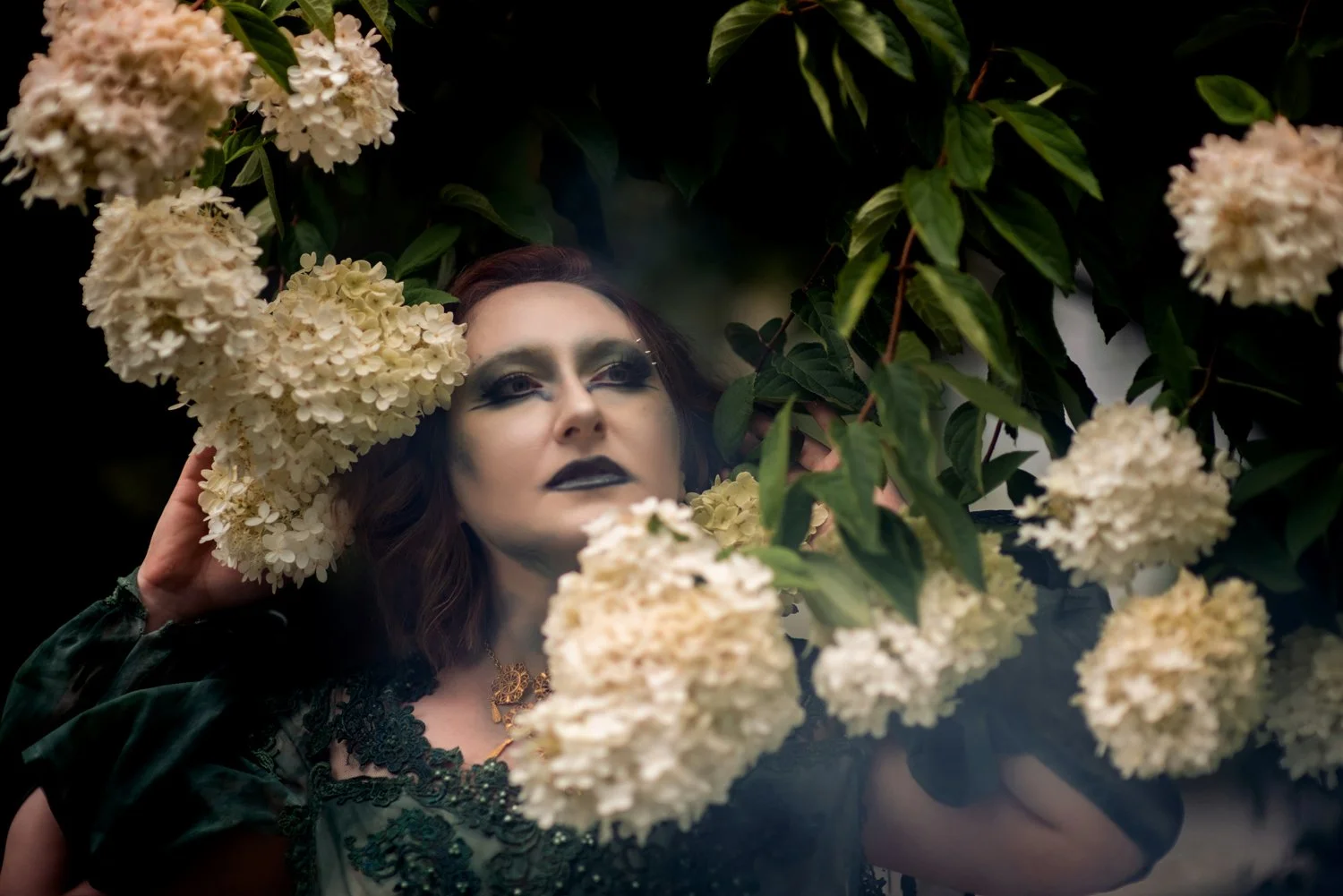 Horror romance author: woman with dark green makeup and dress peering out from around while lilac flowers with dark green leaves