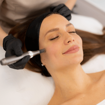 A woman receiving a facial treatment with a cosmetic pen near her cheek, lying down with her eyes closed and a black headband.