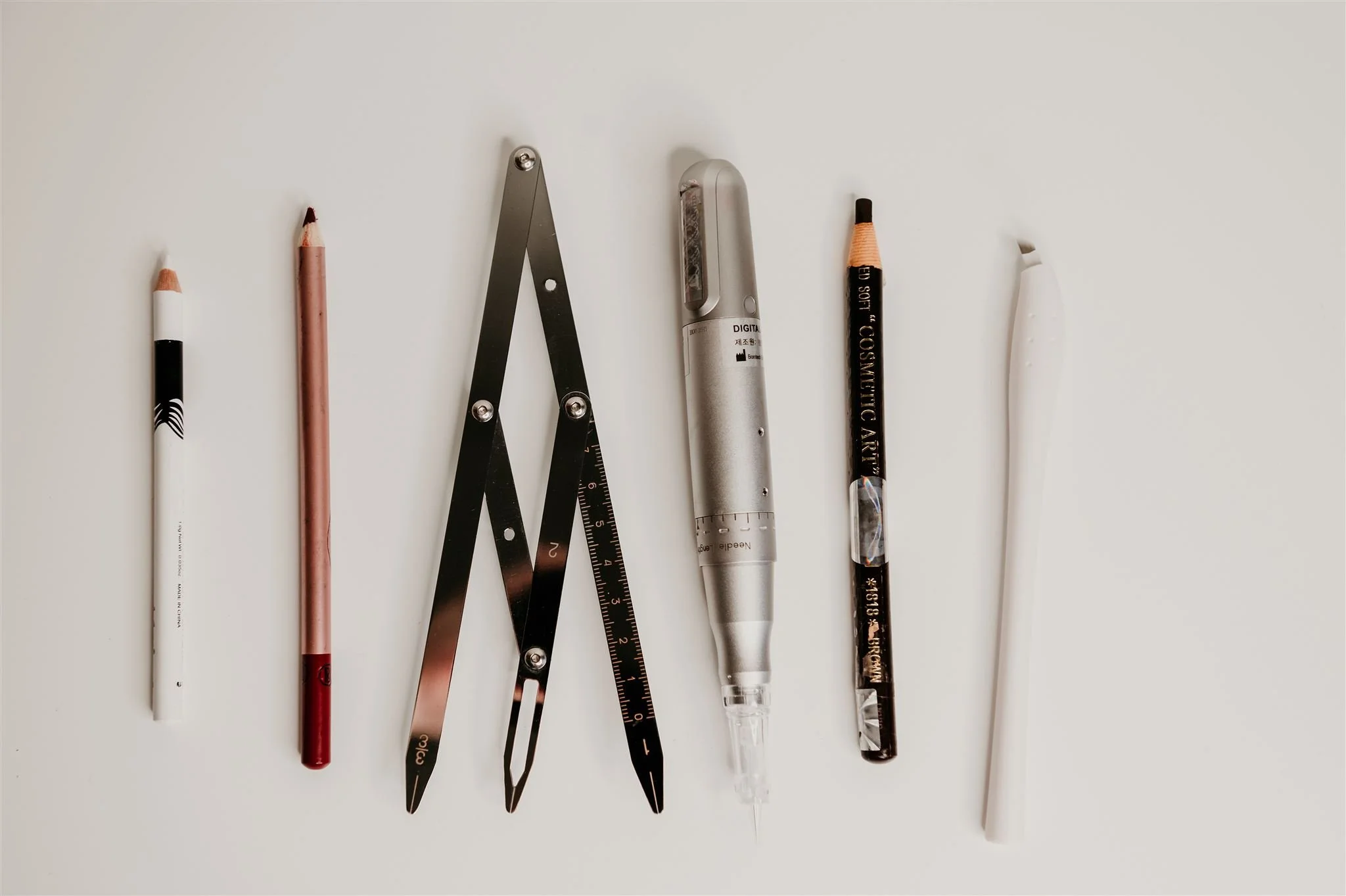 A variety of stationery items including colored pencils, a metal compass, scissors, a silver metallic marker, a black pen, and a white pen are arranged on a plain white surface.