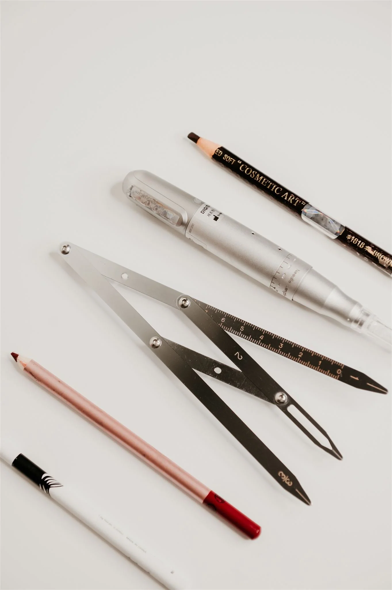 A set of cosmetic and stationery tools including a black eye pencil with gold lettering, a white and silver rollerball pen, a metallic silver ruler, a pink mechanical pencil, and a white marker, all arranged on a white surface.
