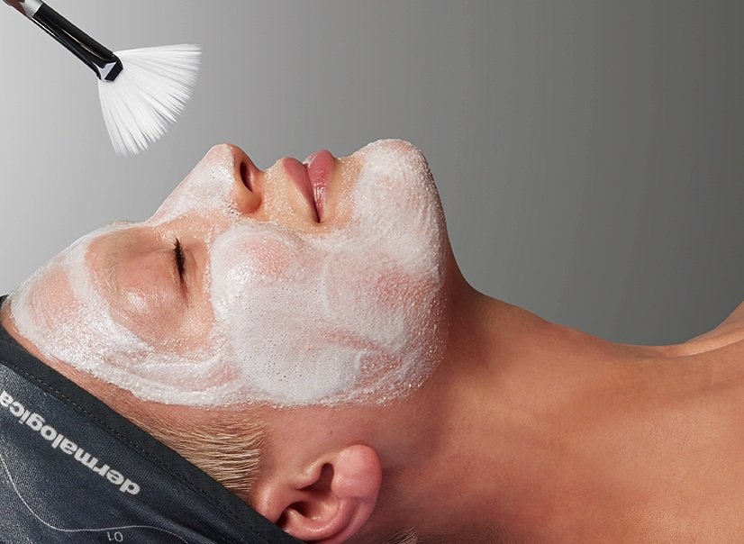 A woman lying on her back with her eyes closed, receiving a facial cleansing treatment with foam or cleanser while a brush applies a product to her face.