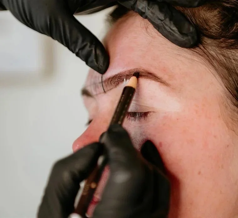 A person with fair skin and light freckles receives an eyebrow tattooing procedure from a professional wearing black gloves, using a tattooing tool on their eyebrow.
