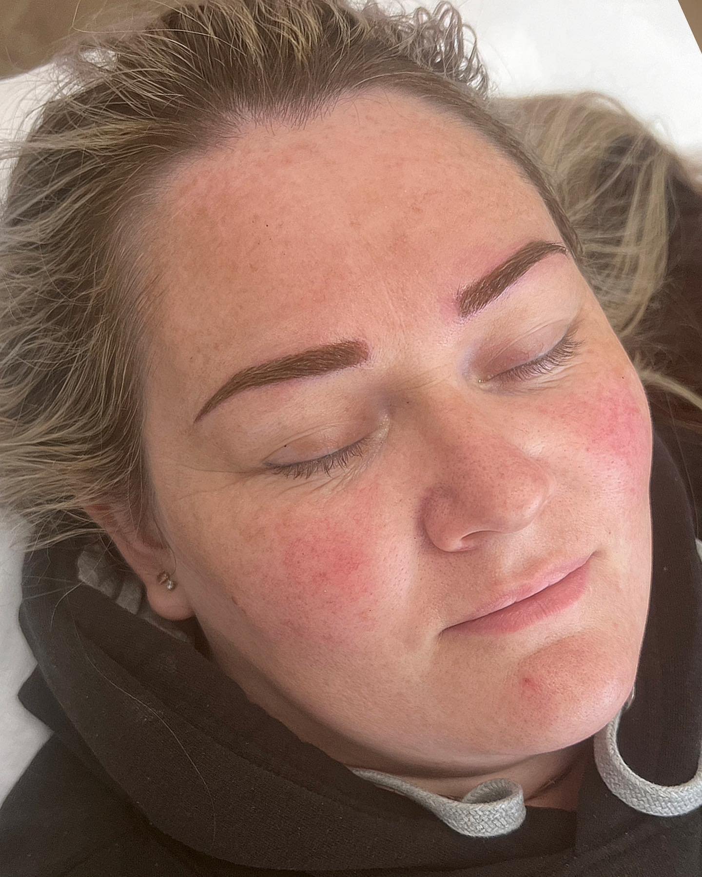 A close-up of a woman's face with freshly done eyebrows, closed eyes, and lightly flushed cheeks, lying on a bed or reclining surface.