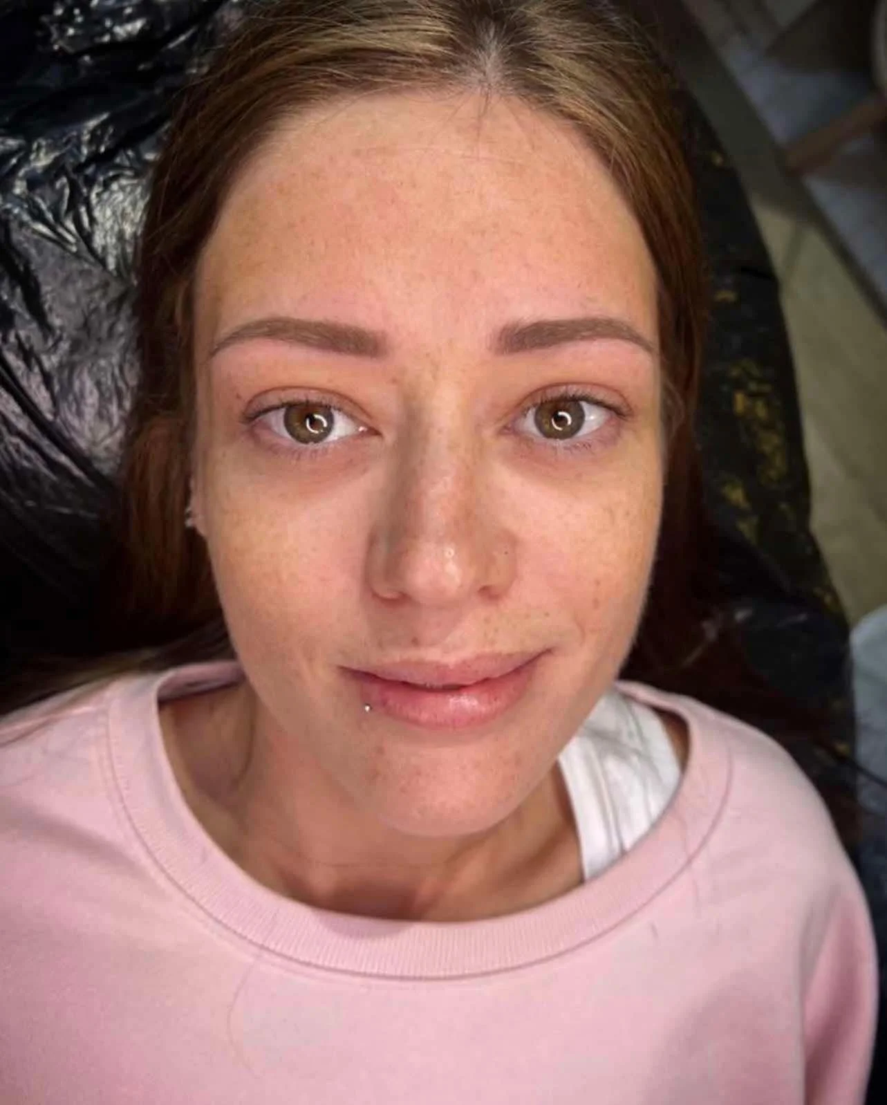 A woman with light brown hair lying on her back, looking up at the camera, wearing a pink shirt, with a slight smile and a dangling lip piercing.