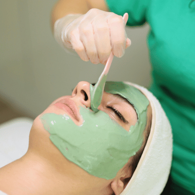 A woman receives a green facial mask at a spa, lying with eyes closed, while a person applies the mask with a brush.