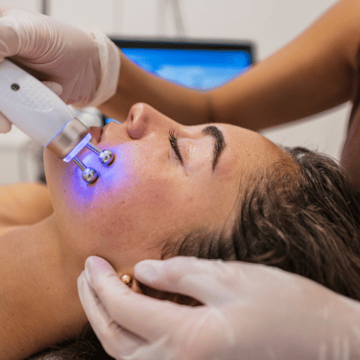 A woman receiving a facial treatment with a device emitting blue light, while lying down with her eyes closed.