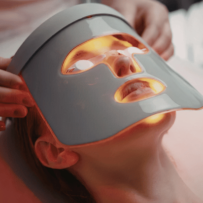 Person receiving a facial treatment with a heated, light-emitting mask while lying down.