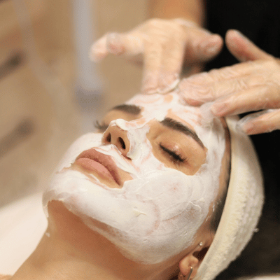Woman receiving a facial treatment with a facial mask applied, while a skincare professional applies another product.