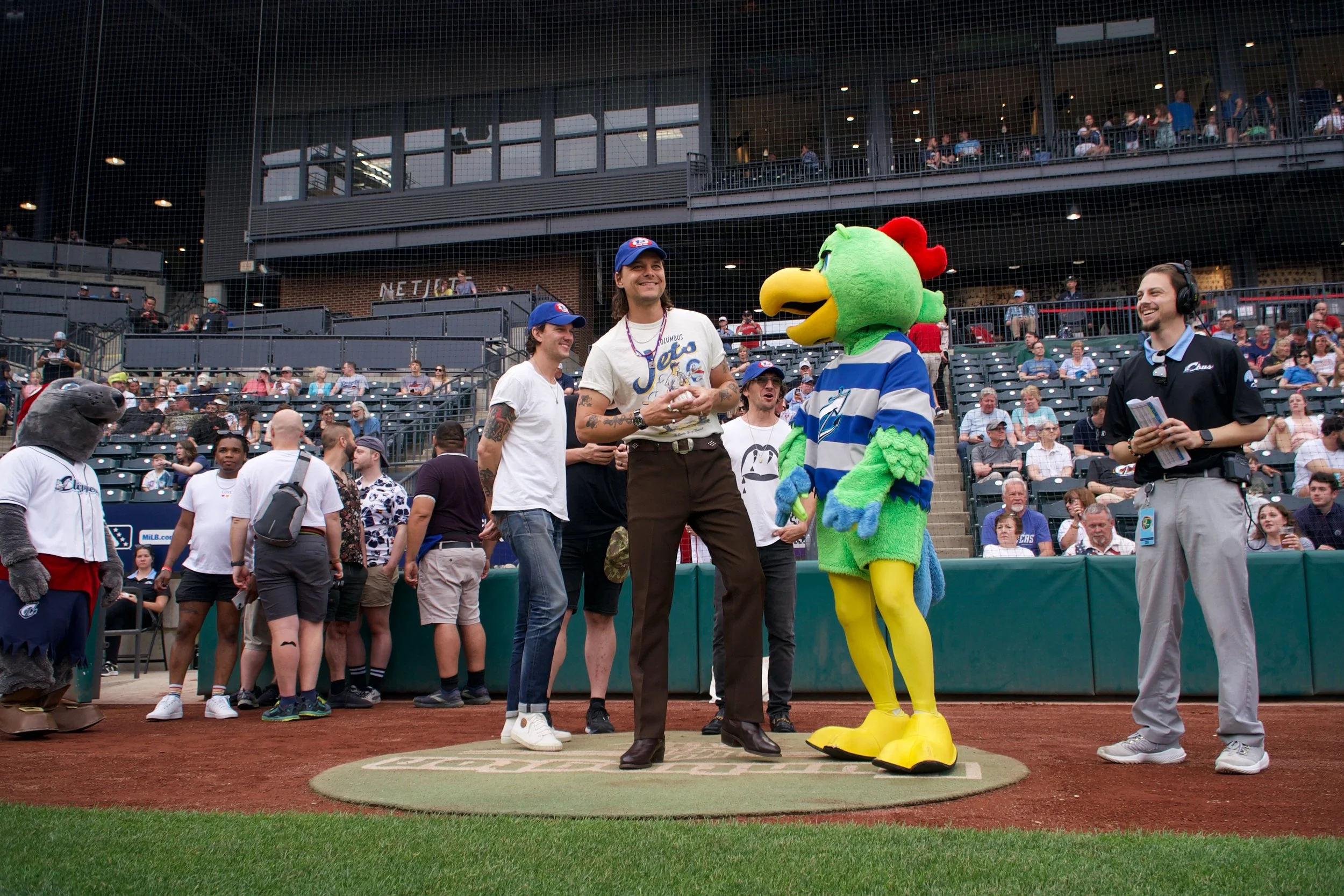 Baseball stadium scene with sports mascot in chicken costume talking to two men, one in a white T-shirt and jeans, the other in a baseball cap and pants, with spectators in the background.