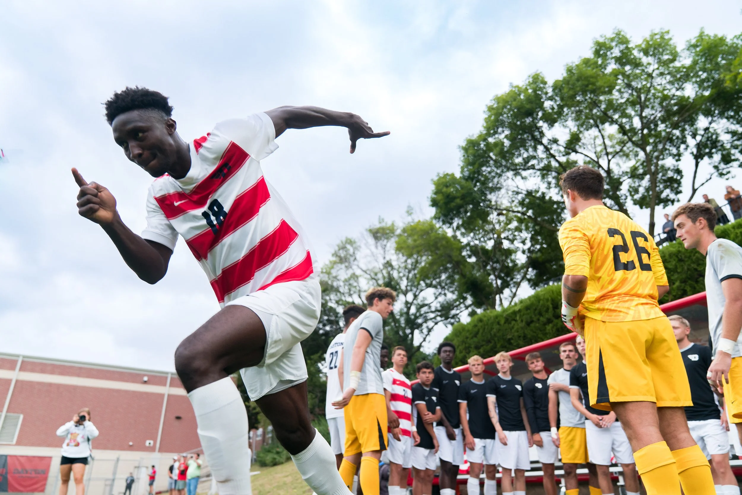 A soccer player in a red and white striped jersey running on the field during a game, with other players and spectators in the background.