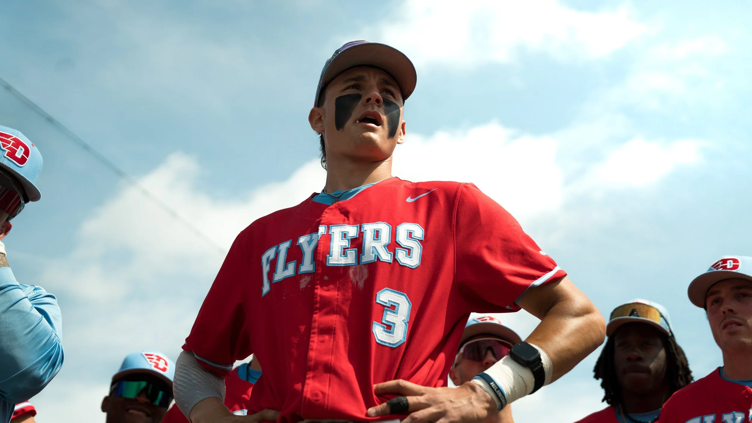 Young male baseball player in a red jersey with 'FLYERS' written on it and number 3, wearing a cap and face paint, standing with teammates during the daytime under a partly cloudy sky.