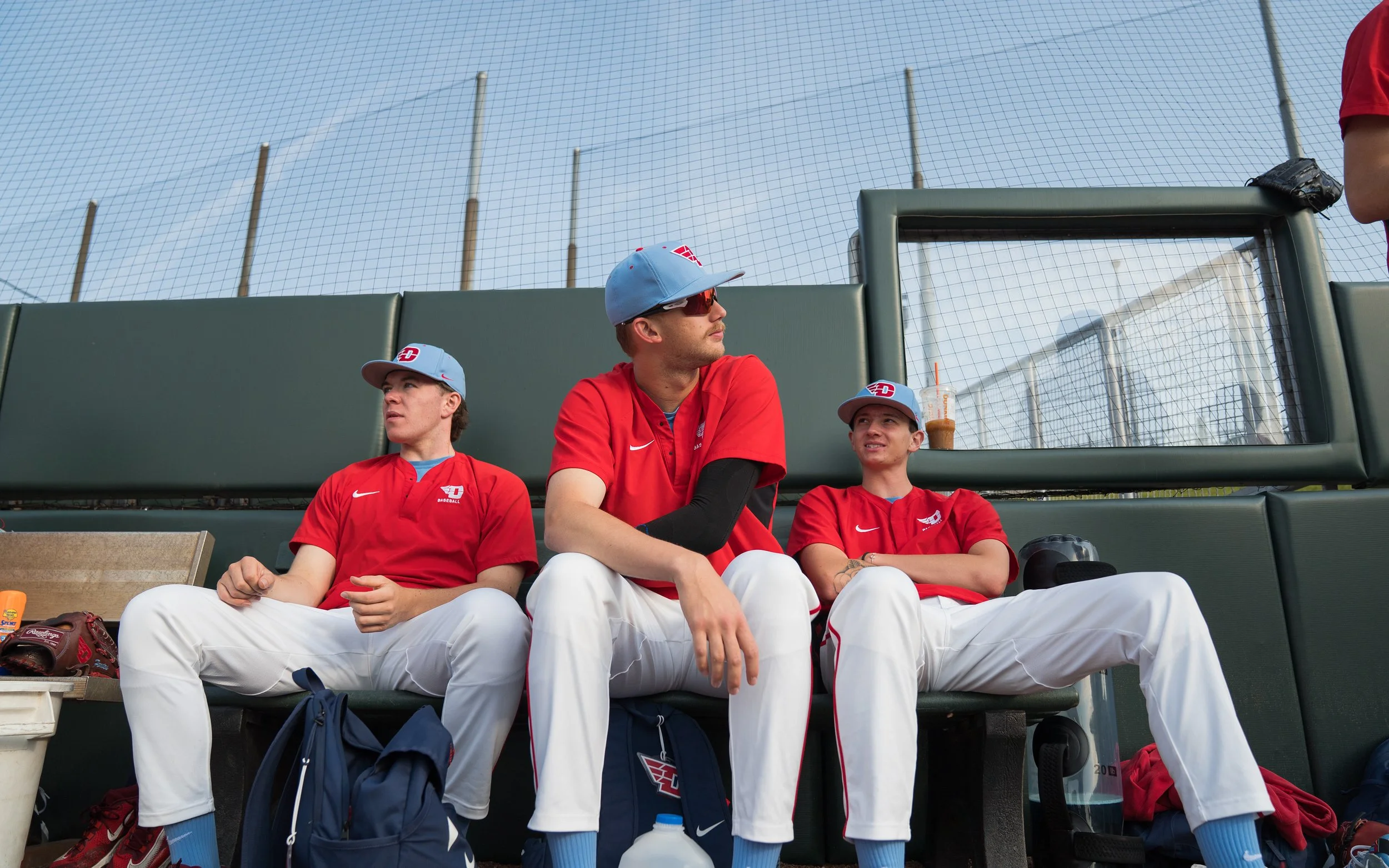 Three baseball players sitting on a dugout bench, wearing red jerseys and white pants, with a baseball glove, water bottles, and a drink in front of them. They are looking in different directions as a chain-link fence and field are visible in the bac