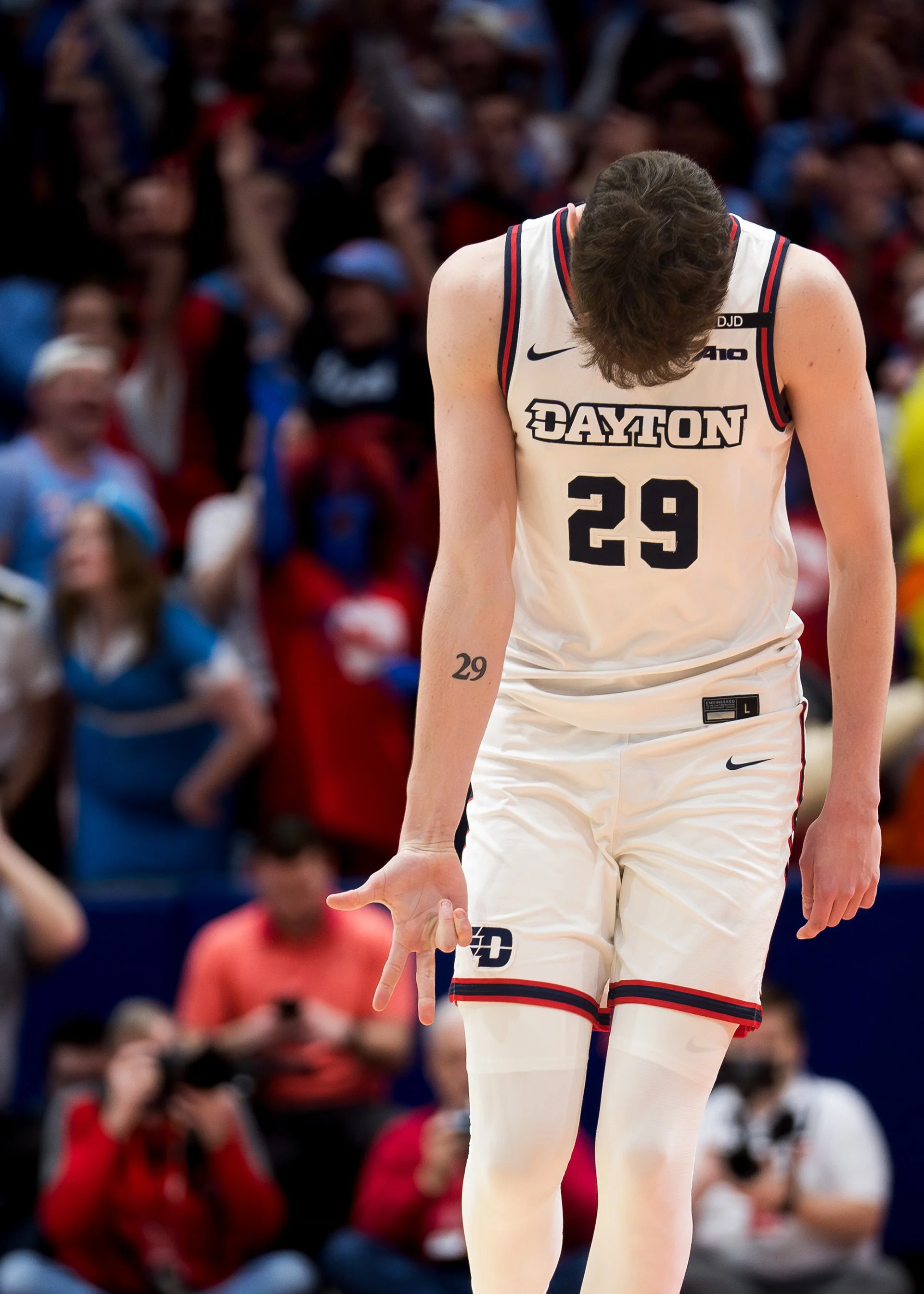 Basketball player wearing a white jersey with 'Dayton' and number 29, making a gesture with their hand during a game, with a blurred crowd in the background.