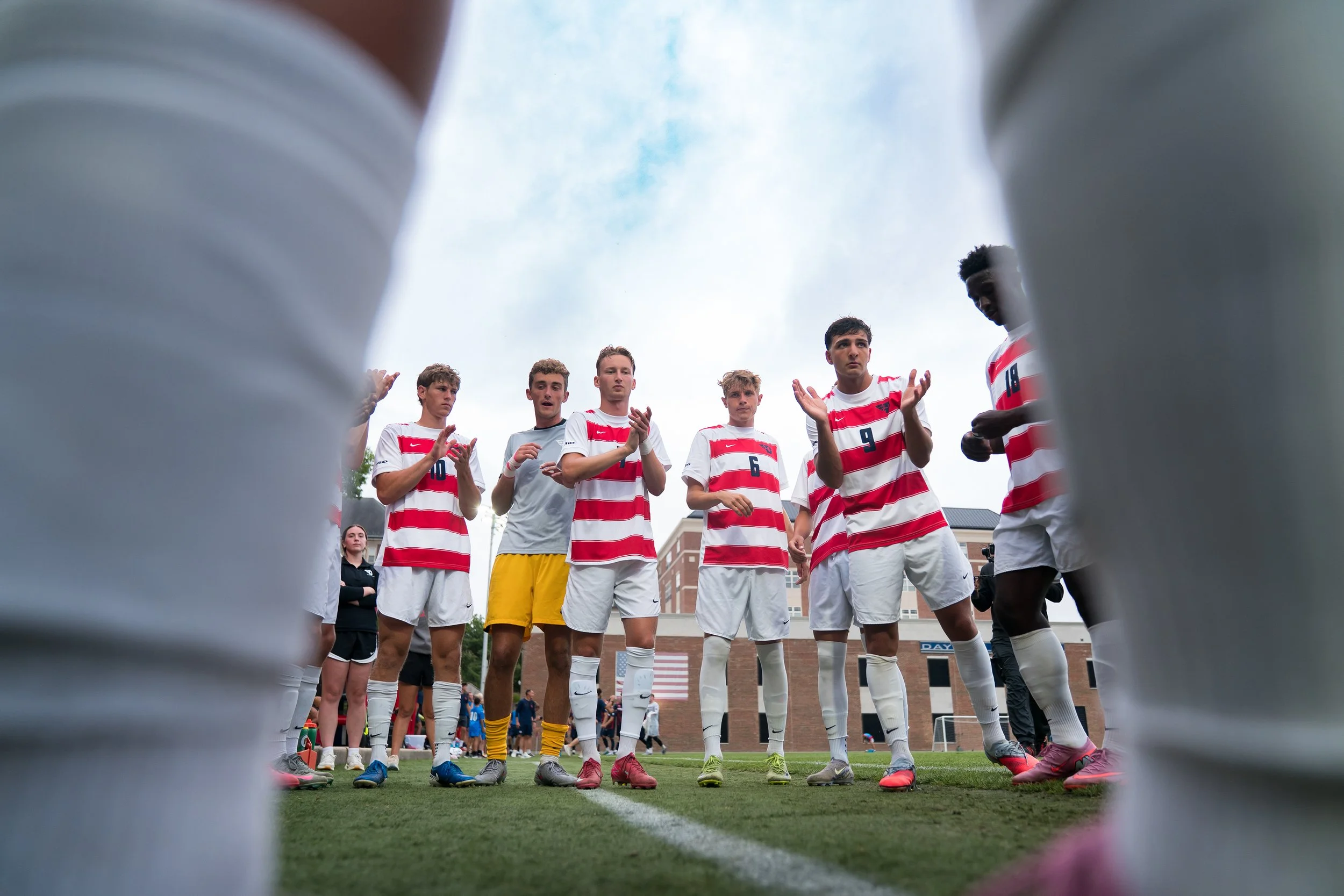 Group of young male soccer players standing in a huddle on a soccer field, seen from a low angle through a gap between two objects, with a building and trees in the background.