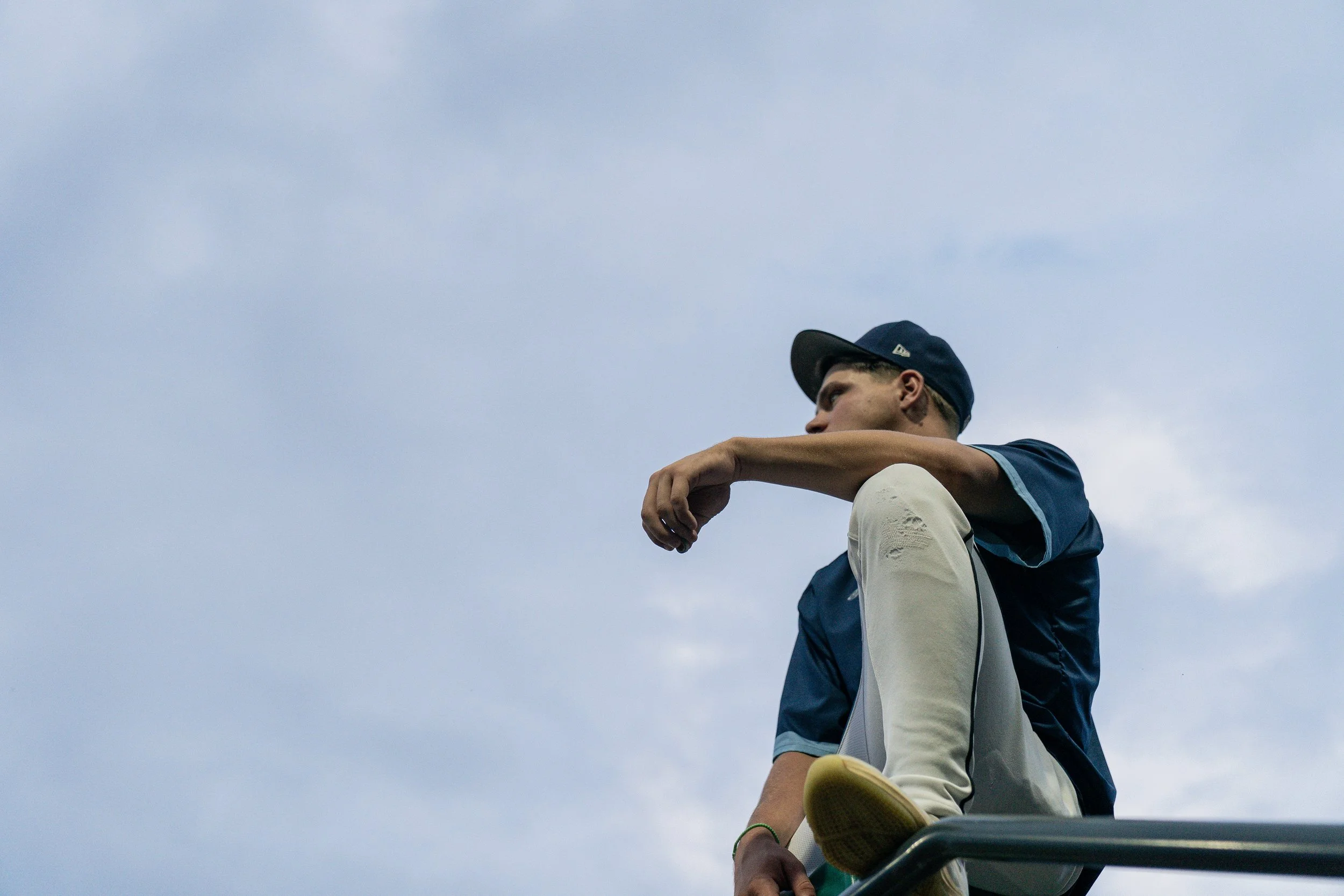 A young man wearing a navy baseball cap and casual clothing sitting on a bench or ledge under a cloudy sky.