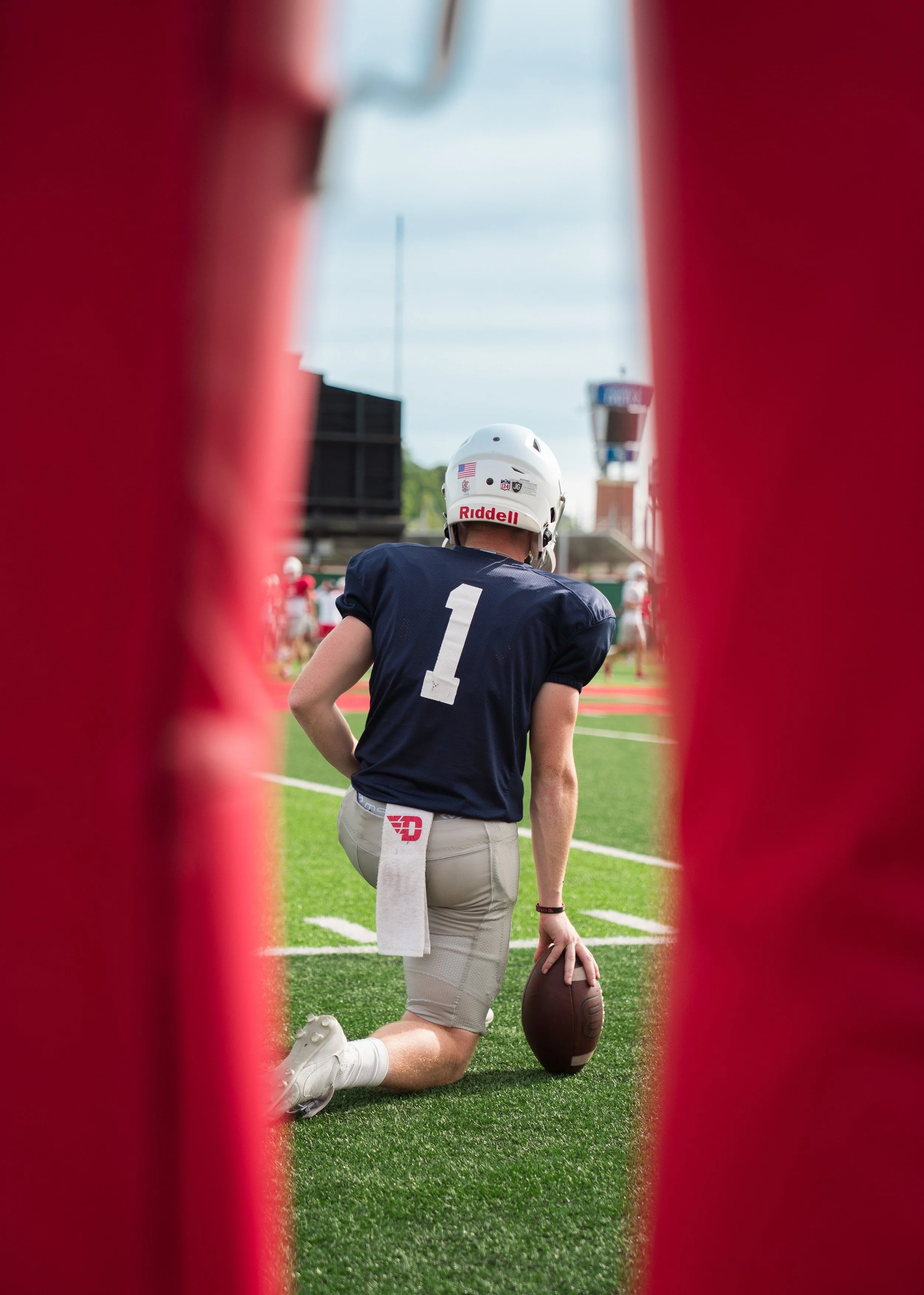 A football player wearing a navy blue uniform with the number 1 on the back, kneeling on a football field, holding a football, viewed through red sideline padding.