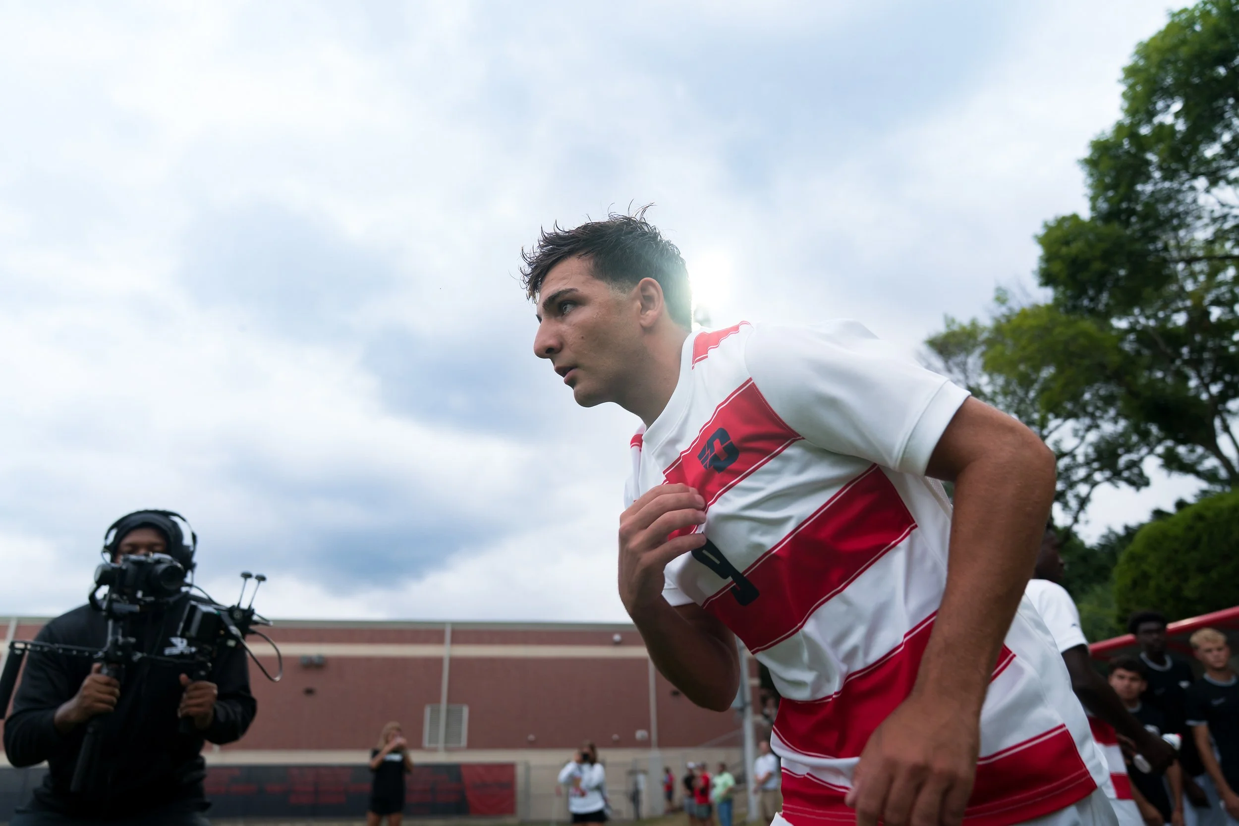 A young man wearing a white and red sports jersey is running outdoors on a cloudy day, with trees and a building in the background. A person with a camera is filming him.