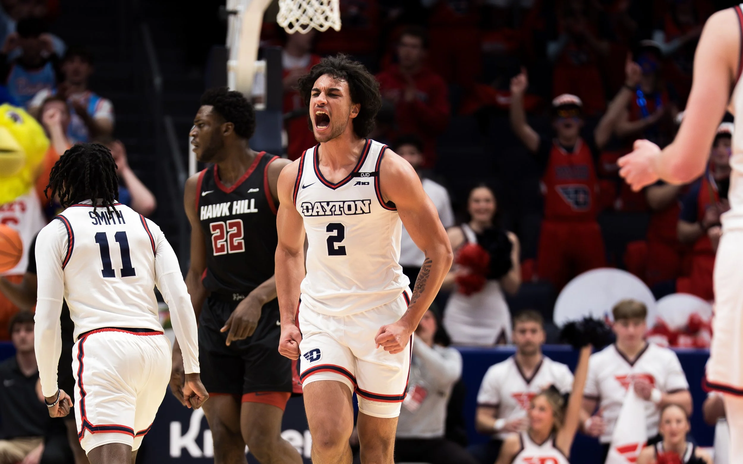 A basketball player wearing a white Dayton jersey with the number 2 is celebrating on the court, surrounded by teammates and opponents, with crowd members and cheerleaders visible in the background.