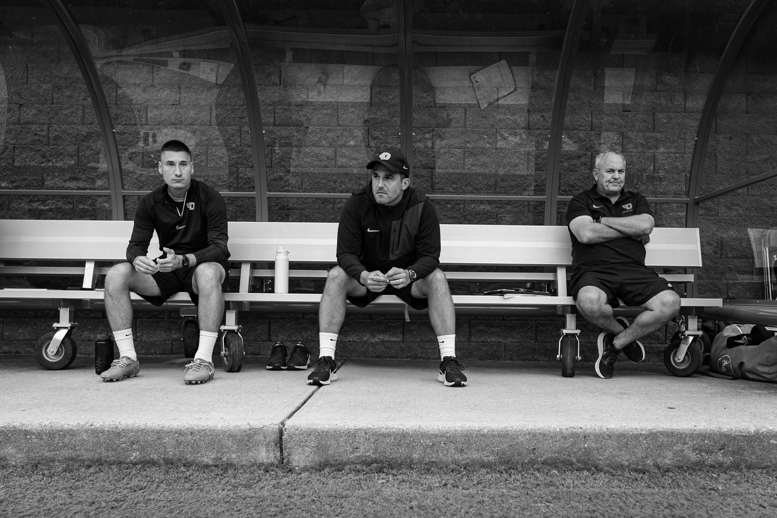 Three men sitting on a bench inside a glass-walled shelter against a brick wall, one on the left in athletic gear, one in the middle with a water bottle, and one on the right with crossed arms and a backpack on the ground.