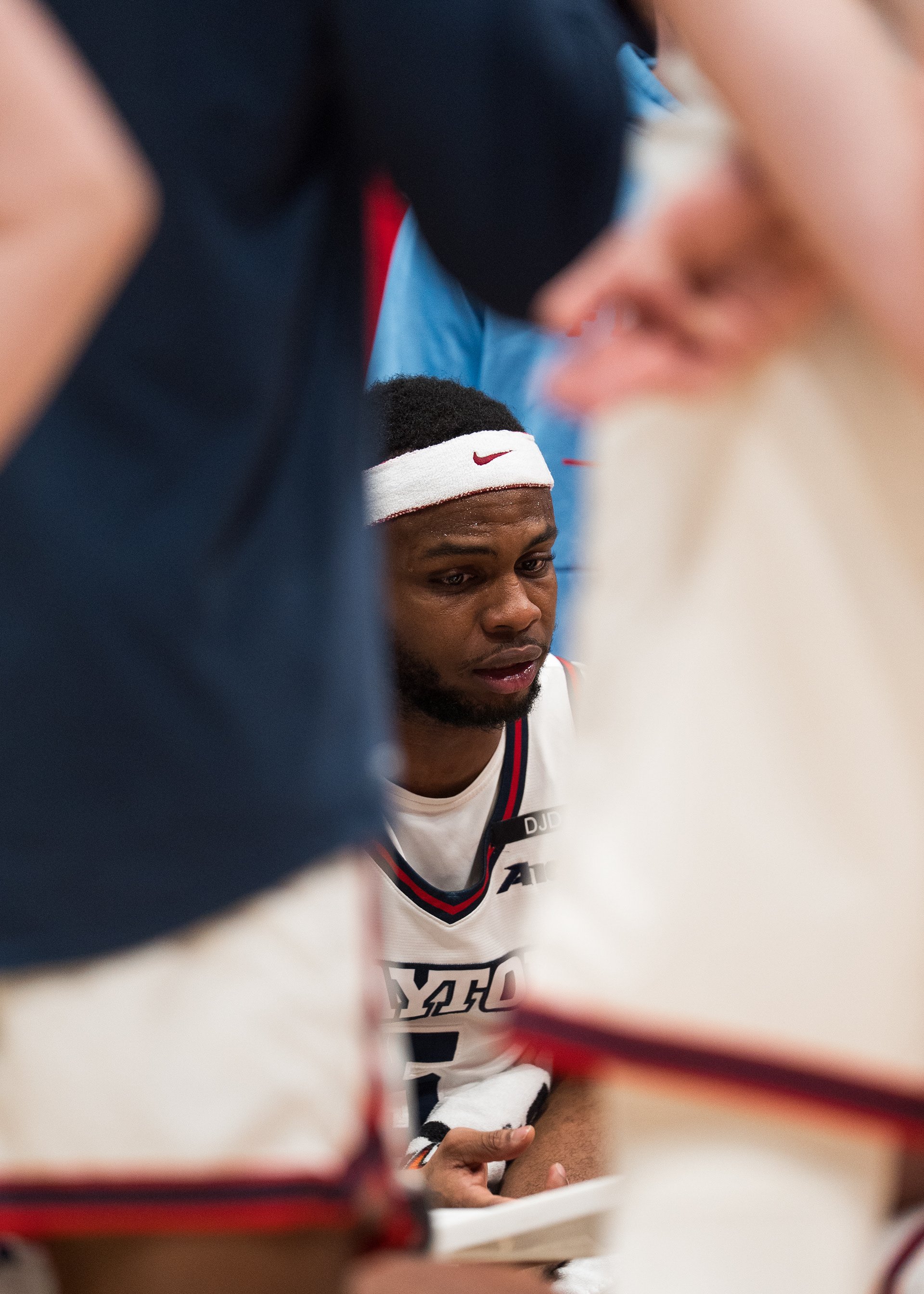 Close-up of a basketball player with a headband, wearing a white jersey, sitting on the bench surrounded by teammates and staff.