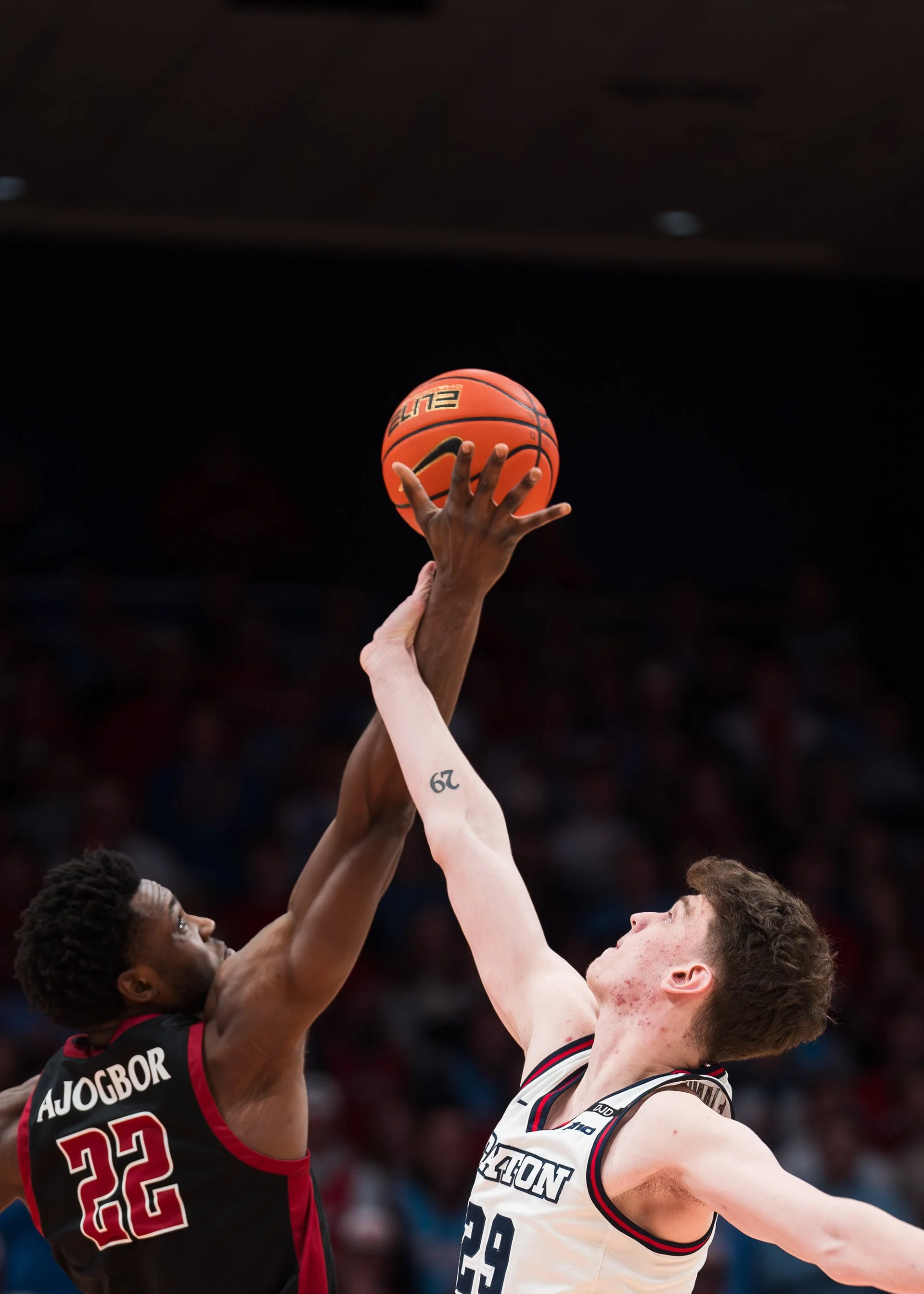 Two basketball players, one with a dark skin tone and the other with a light skin tone, are reaching for a basketball during a game. The player with dark skin is wearing a black jersey with red and white accents and the number 22. The player with lig