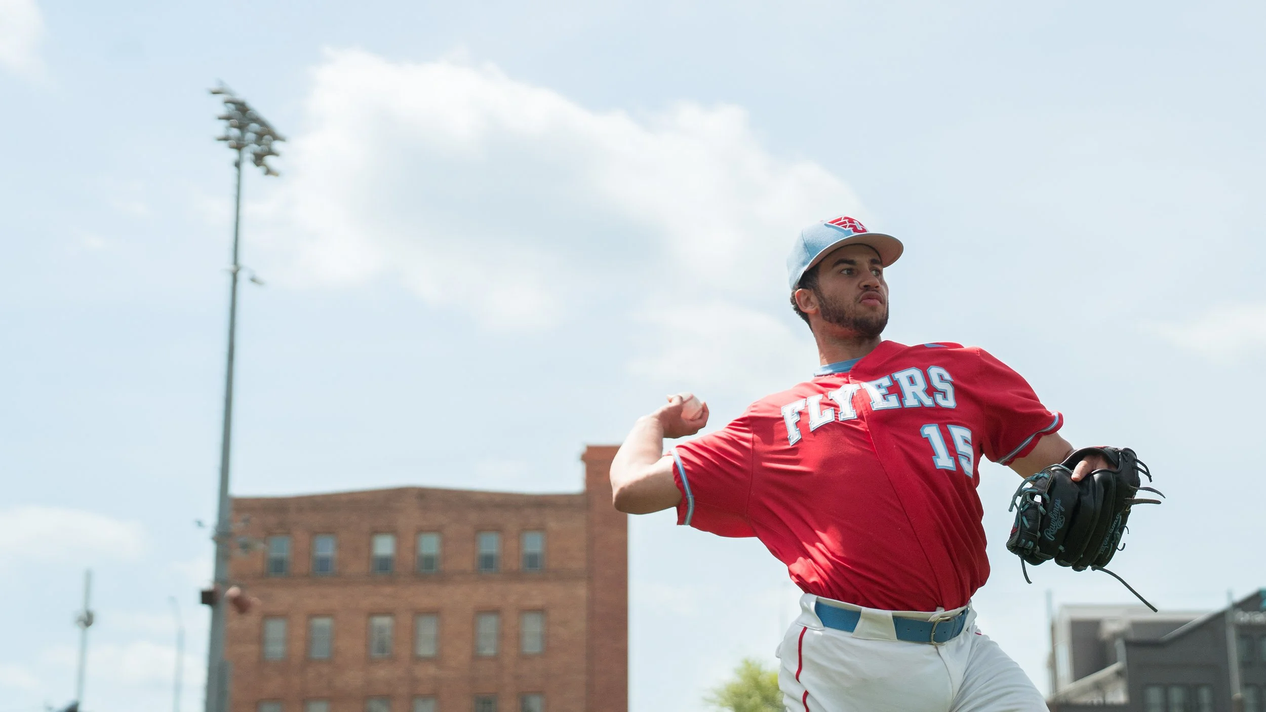 A baseball player wearing a red jersey and gray cap with a red logo, appears to be pitching a ball during a game outdoors on a partly cloudy day, with a tall floodlight and a brick building in the background.