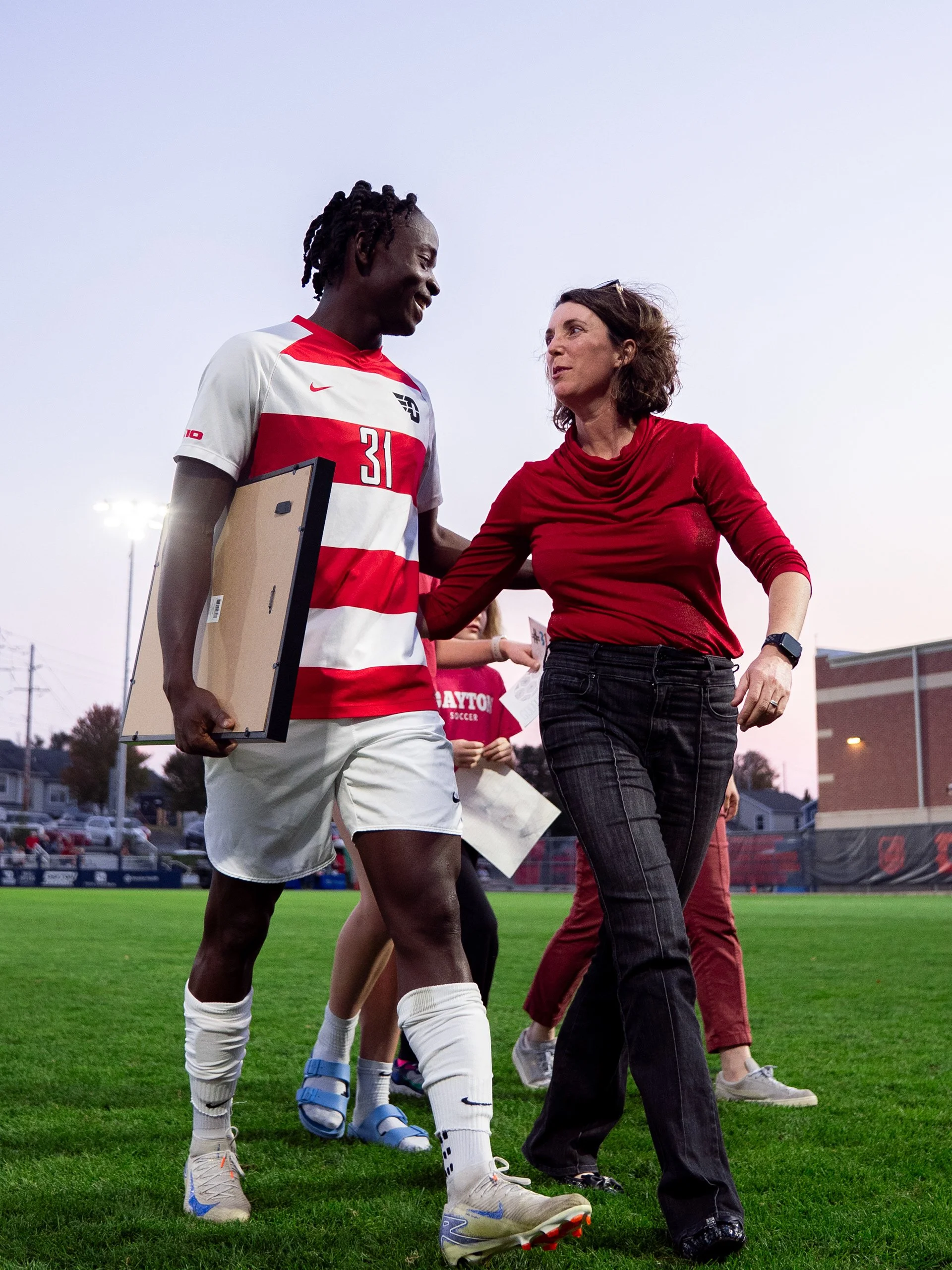 A soccer player in a red and white uniform talking to a woman in a red long-sleeve shirt, on a soccer field during evening hours.