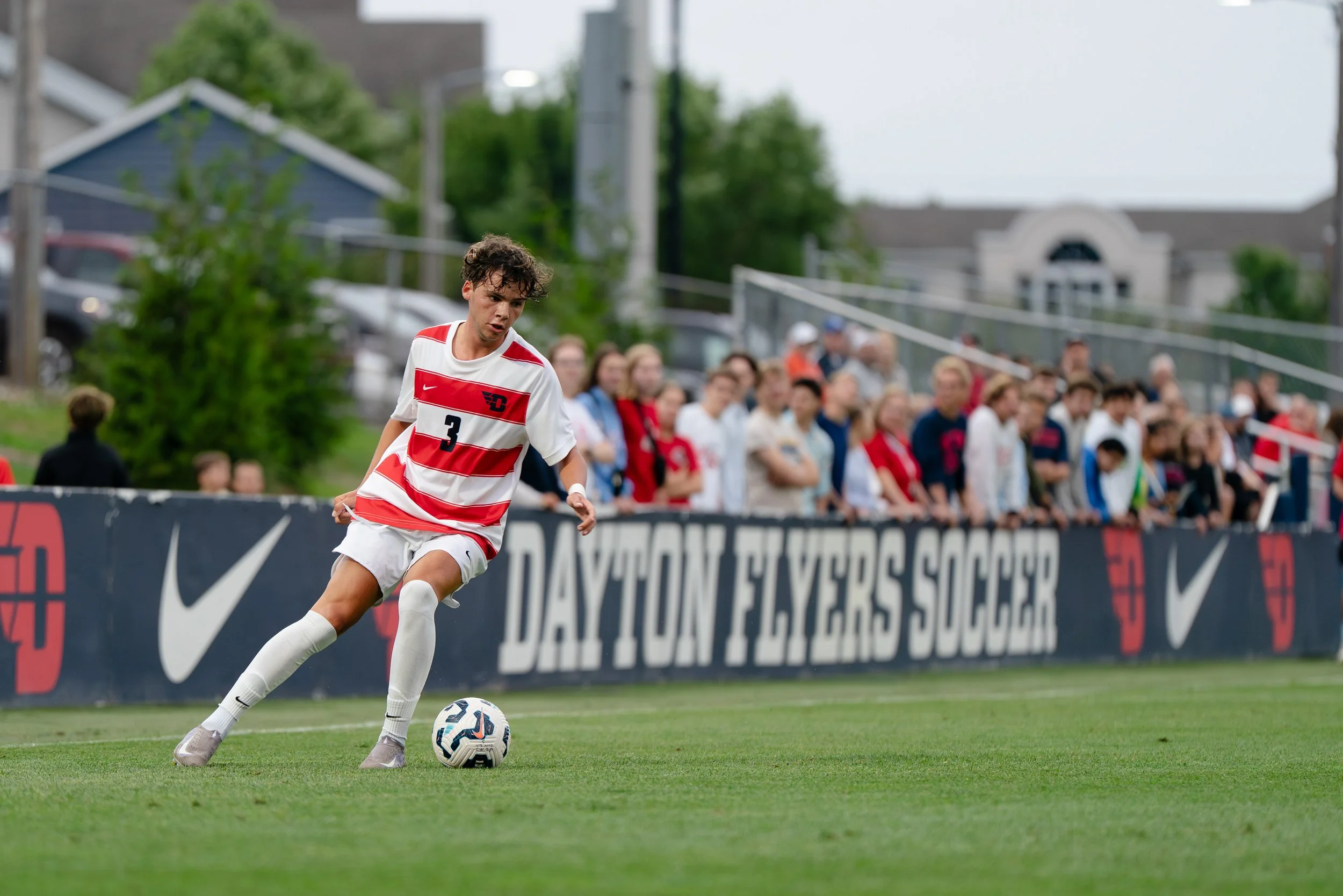 A young male soccer player in a red and white striped jersey with the number 3, white shorts, and cleats, is about to kick a soccer ball on a green field during a match. Spectators and buildings are visible in the background.