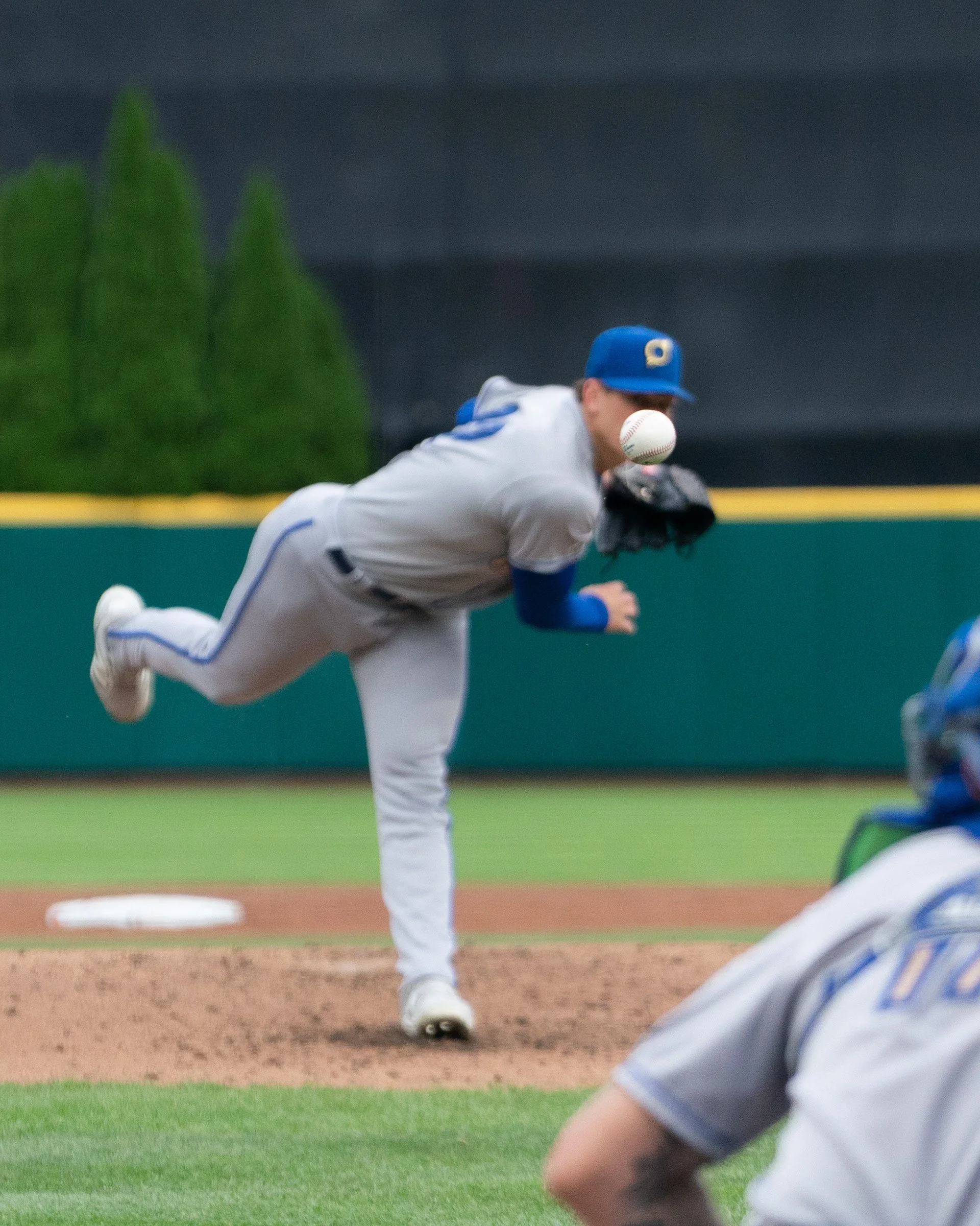A baseball player in a gray uniform and blue cap is pitching, with a baseball close to his face, on a baseball field.