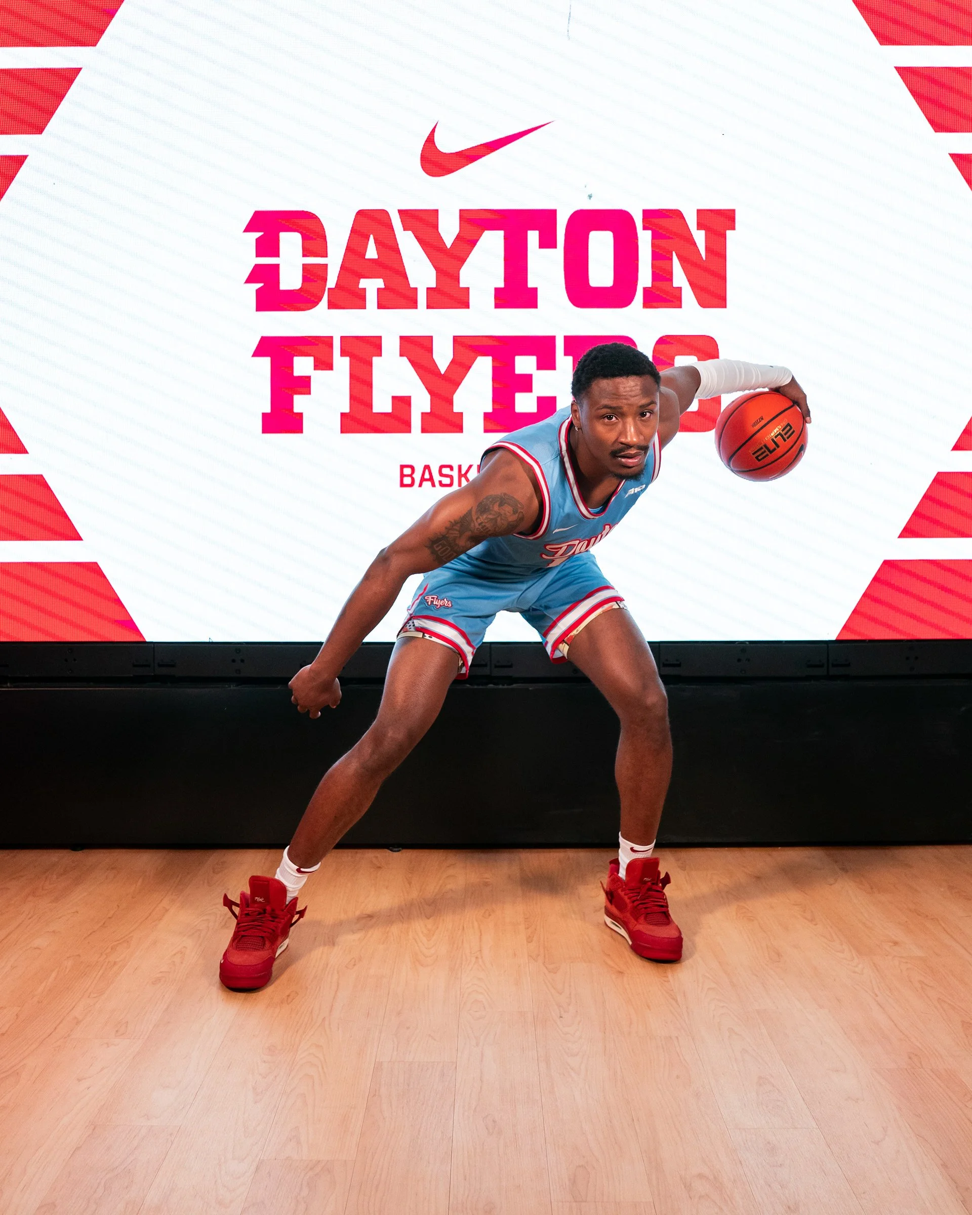 An African-American man playing basketball in front of a digital screen that says 'Dayton Flyers' and features a Nike logo. He is wearing a blue basketball uniform with red and white accents and red athletic shoes. He is holding a basketball in his r
