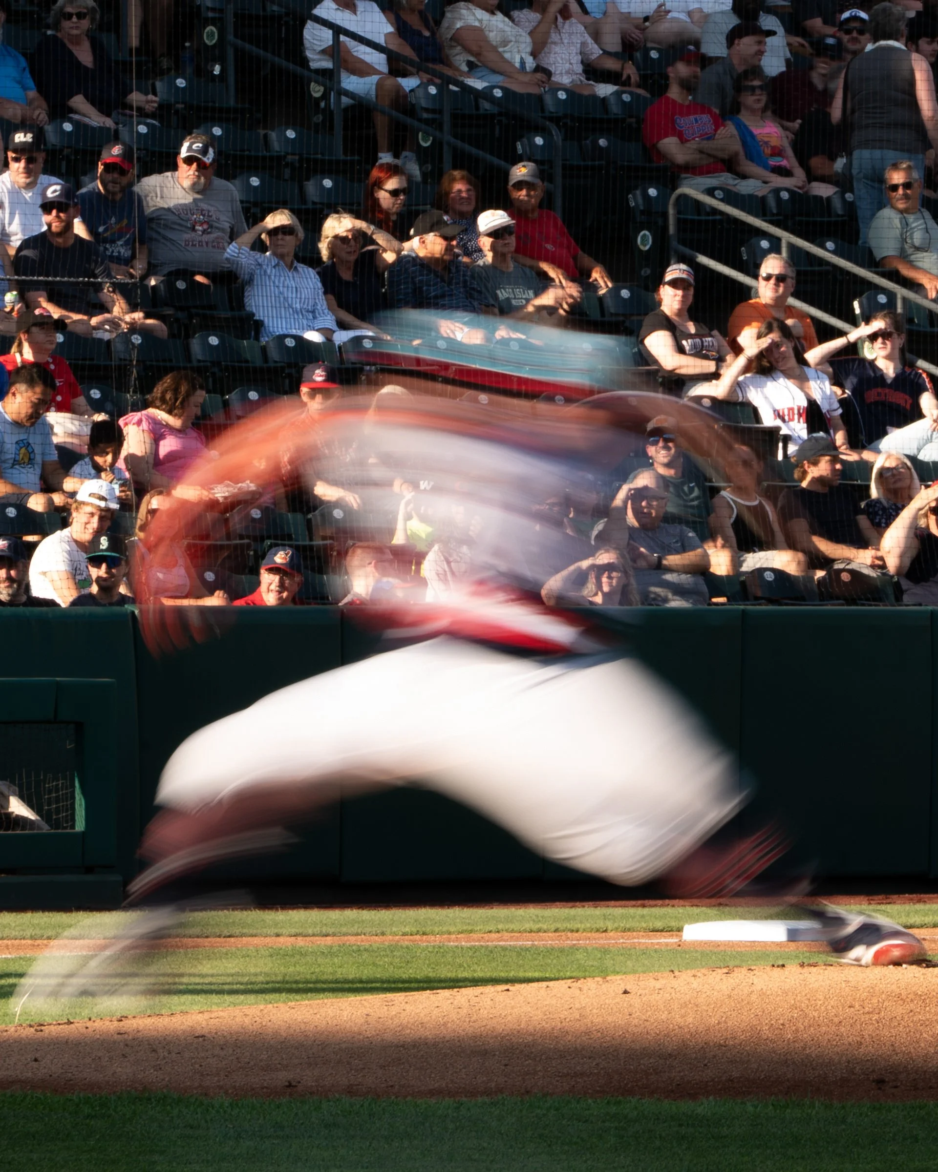 A baseball field with an approaching player blurred from motion, and spectators sitting in the stands behind a net. The spectators are watching the game, some wearing caps and sunglasses.