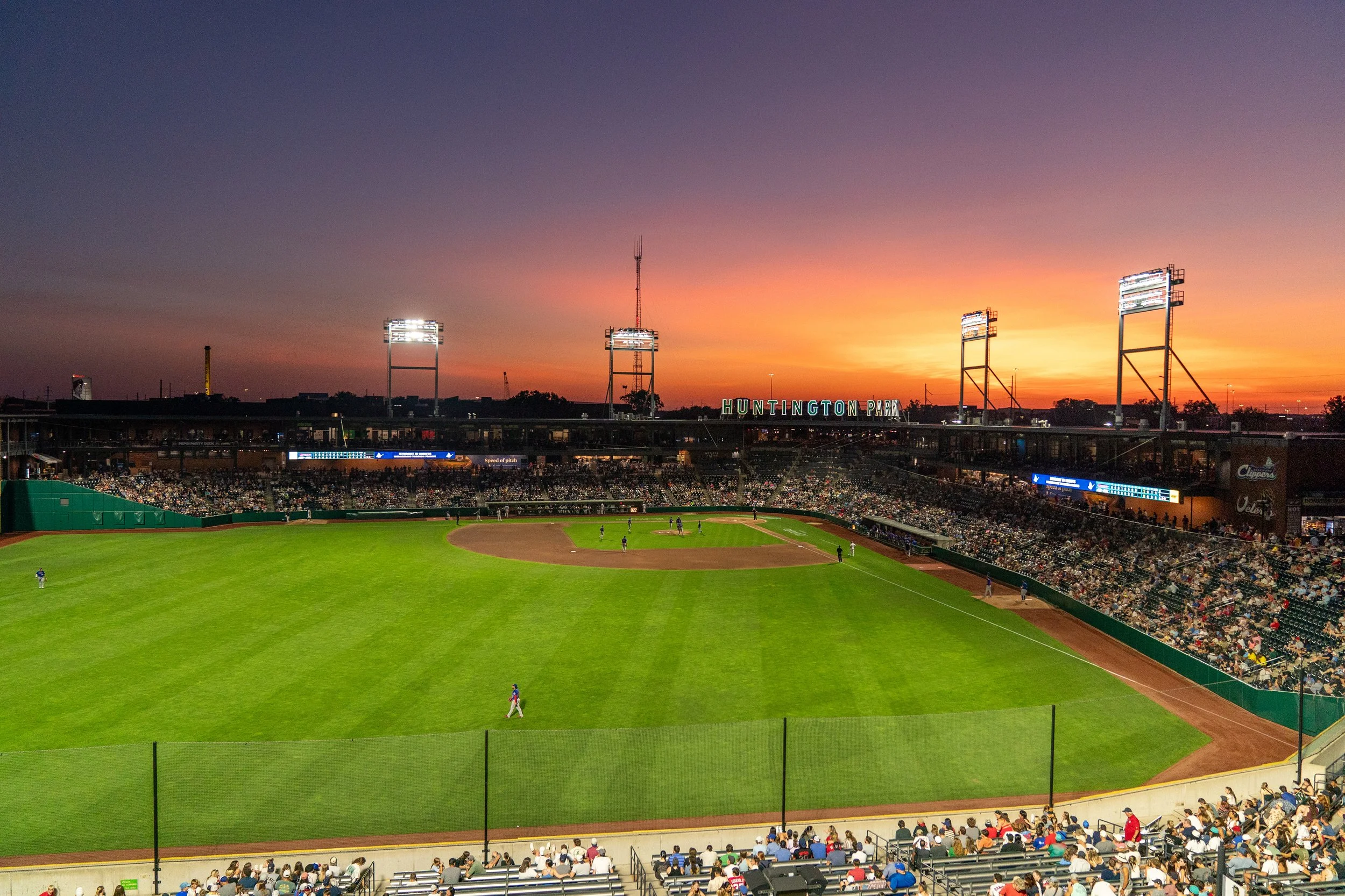 Sunset over a baseball stadium with players on the field and fans in the stands. The sign reads 'Huntington Park' and there are large floodlights and a tall communication tower.