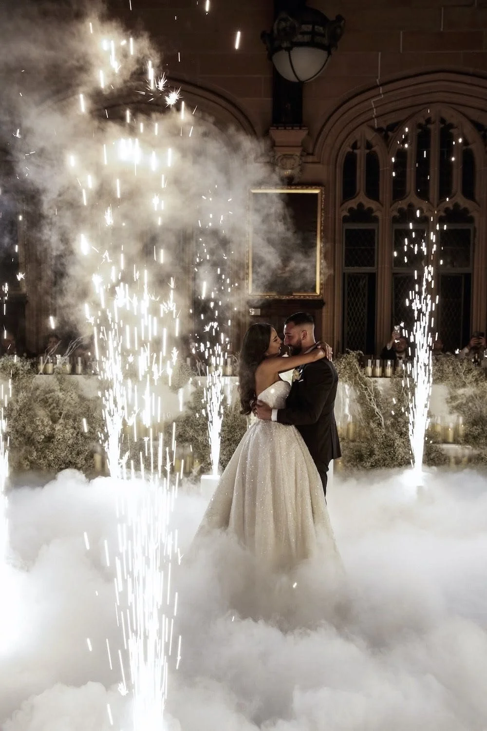 A bride and groom dancing surrounded by fireworks and smoke at their wedding reception in a grand, decorated hall.