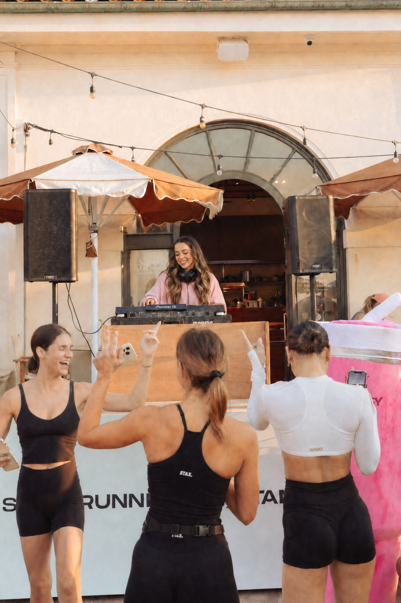A group of women at a fitness event, some wearing black workout clothes and a woman DJ performing with headphones in front of a crowd. Others are with raising hands and interacting.