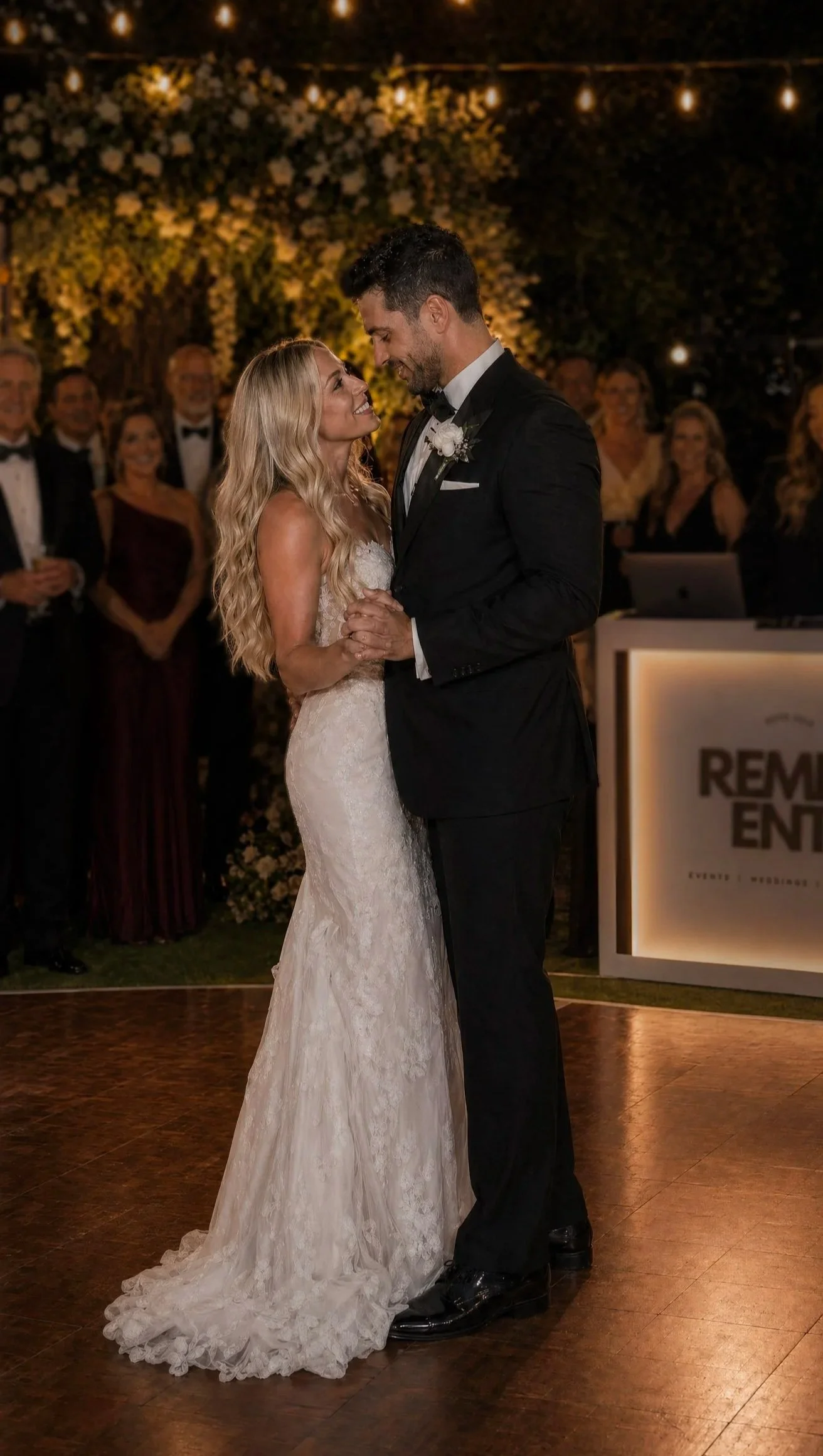 A bride and groom dancing at their wedding reception at night, surrounded by guests, with string lights and floral decorations in the background.