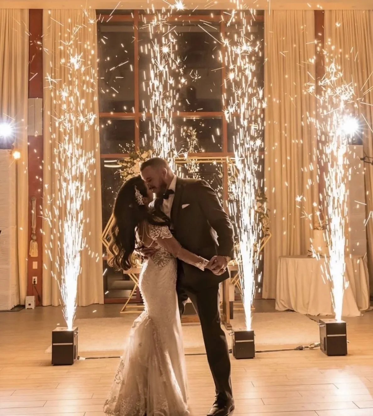A bride and groom dancing together at their wedding reception surrounded by sparklers and fireworks. They are holding hands and sharing a close moment in front of a decorated backdrop with large windows and curtains.