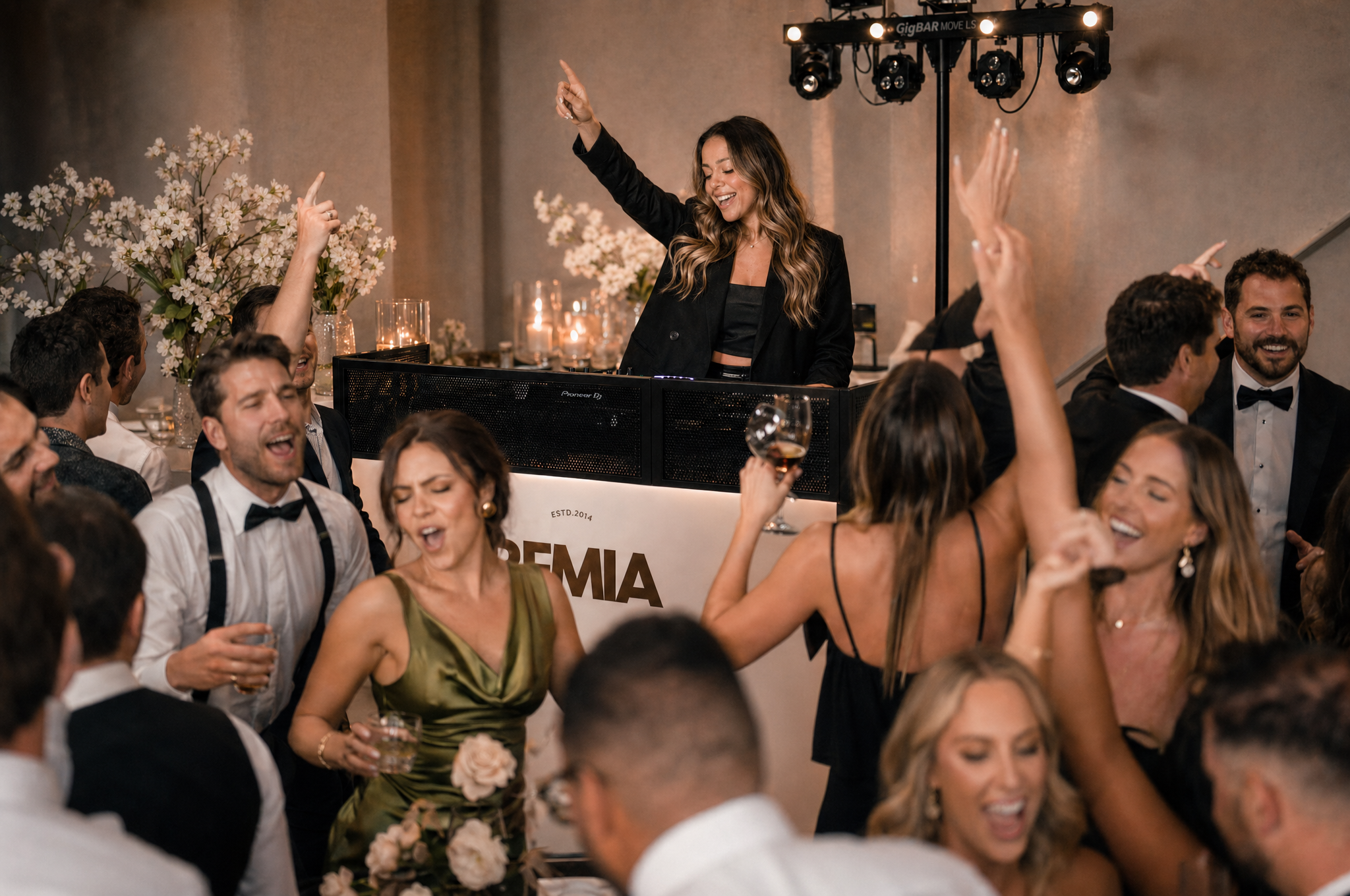 A woman with long wavy hair dancing and smiling behind a DJ booth at a lively party surrounded by guests, some with glasses of wine, in an elegant setting with floral decorations and candles.