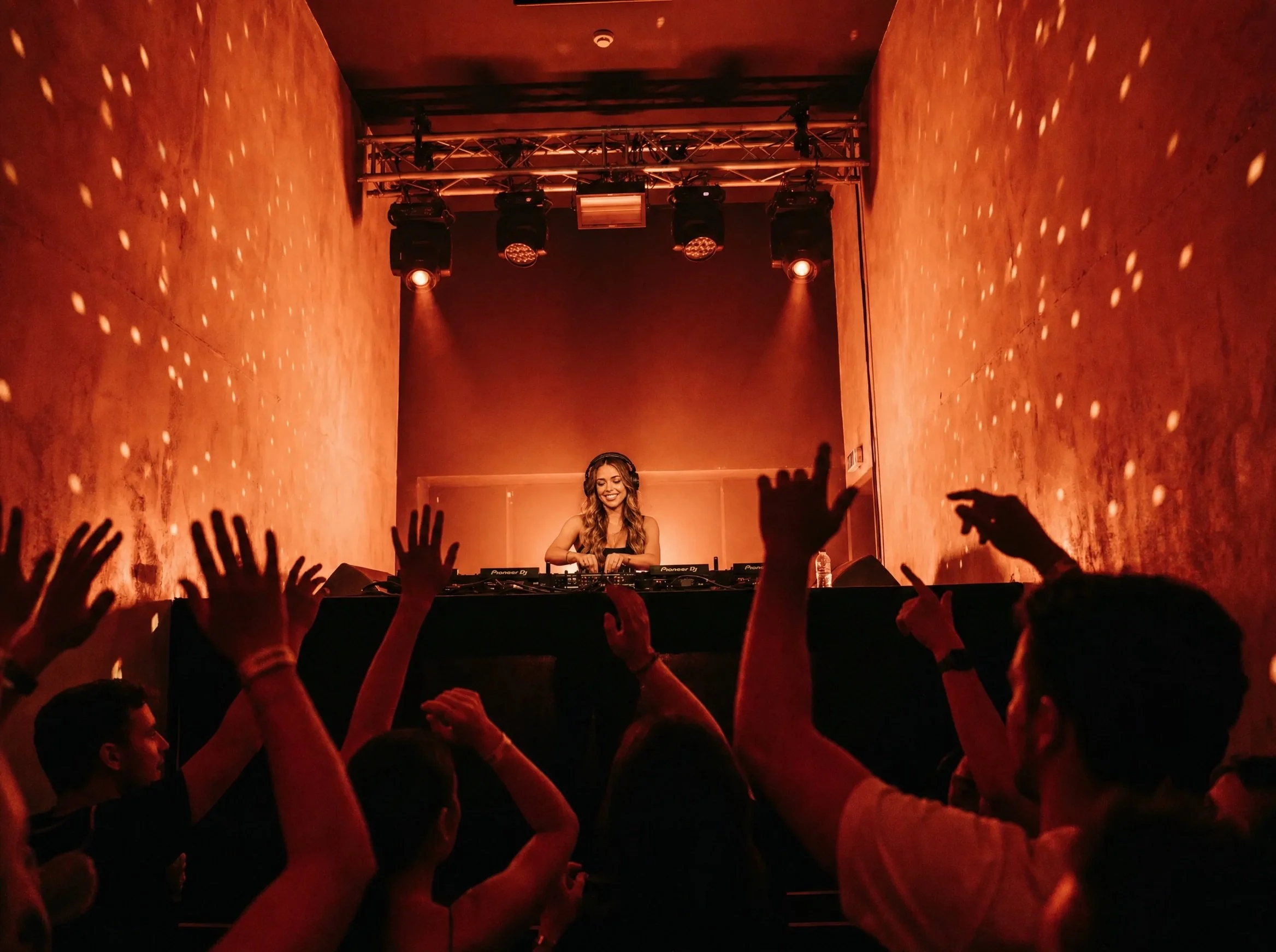 A woman DJ performing on stage at a concert, with hands of audience members reaching towards her in the foreground, the stage is illuminated with orange lighting.