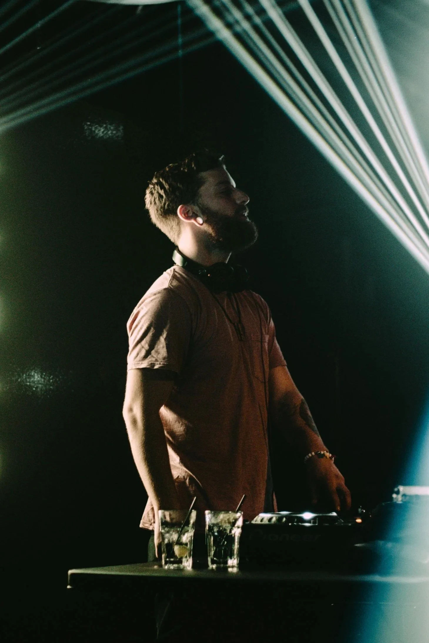 A male DJ with a beard, wearing a brown shirt and headphones around his neck, standing behind a DJ console in a dark setting with colored lights.