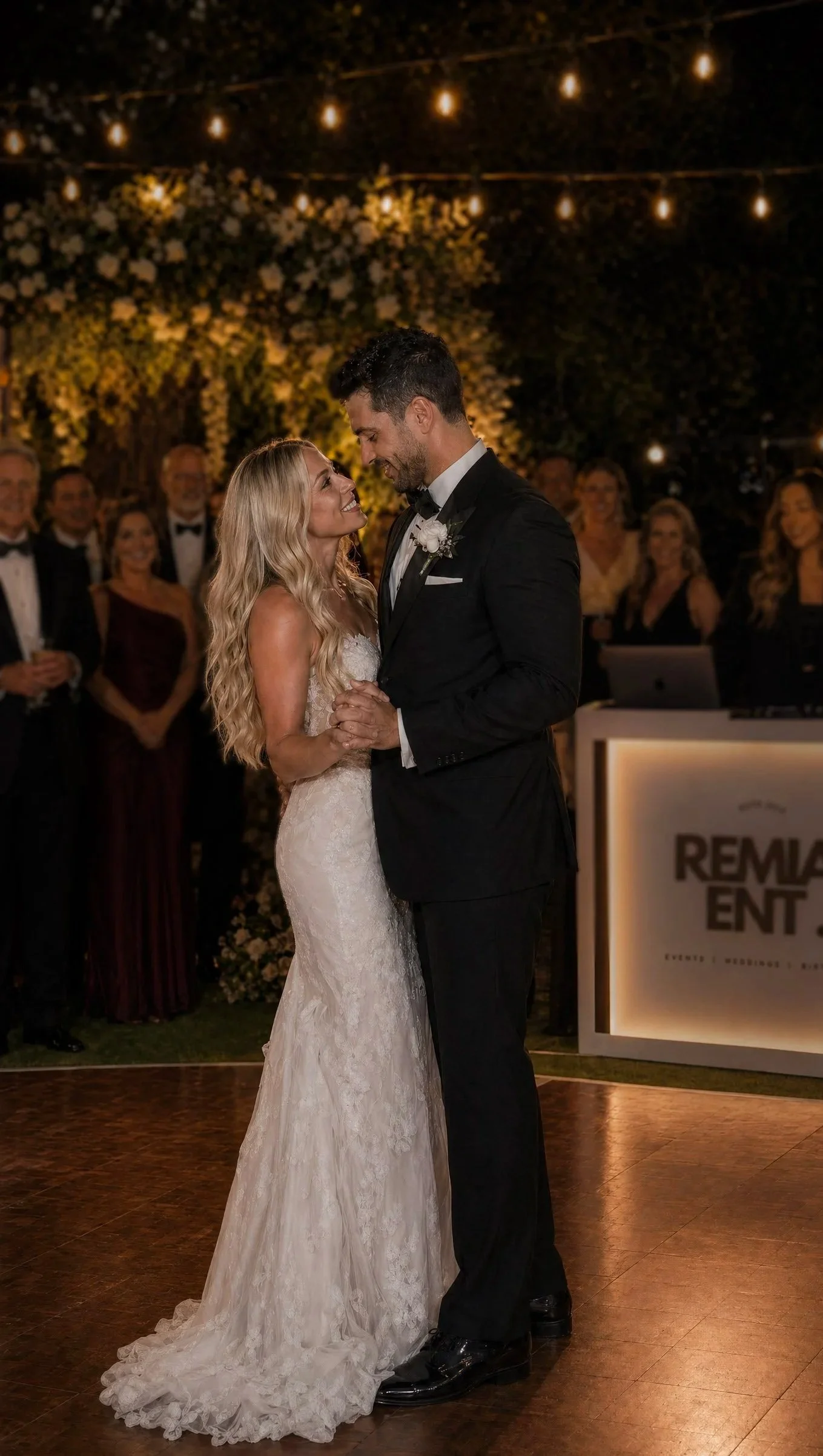 A bride and groom sharing a first dance at their wedding reception at night. The bride is wearing a lace wedding dress, and the groom is in a black tuxedo. They are looking into each other's eyes, smiling. In the background, guests are watching, and there are string lights and floral decorations.