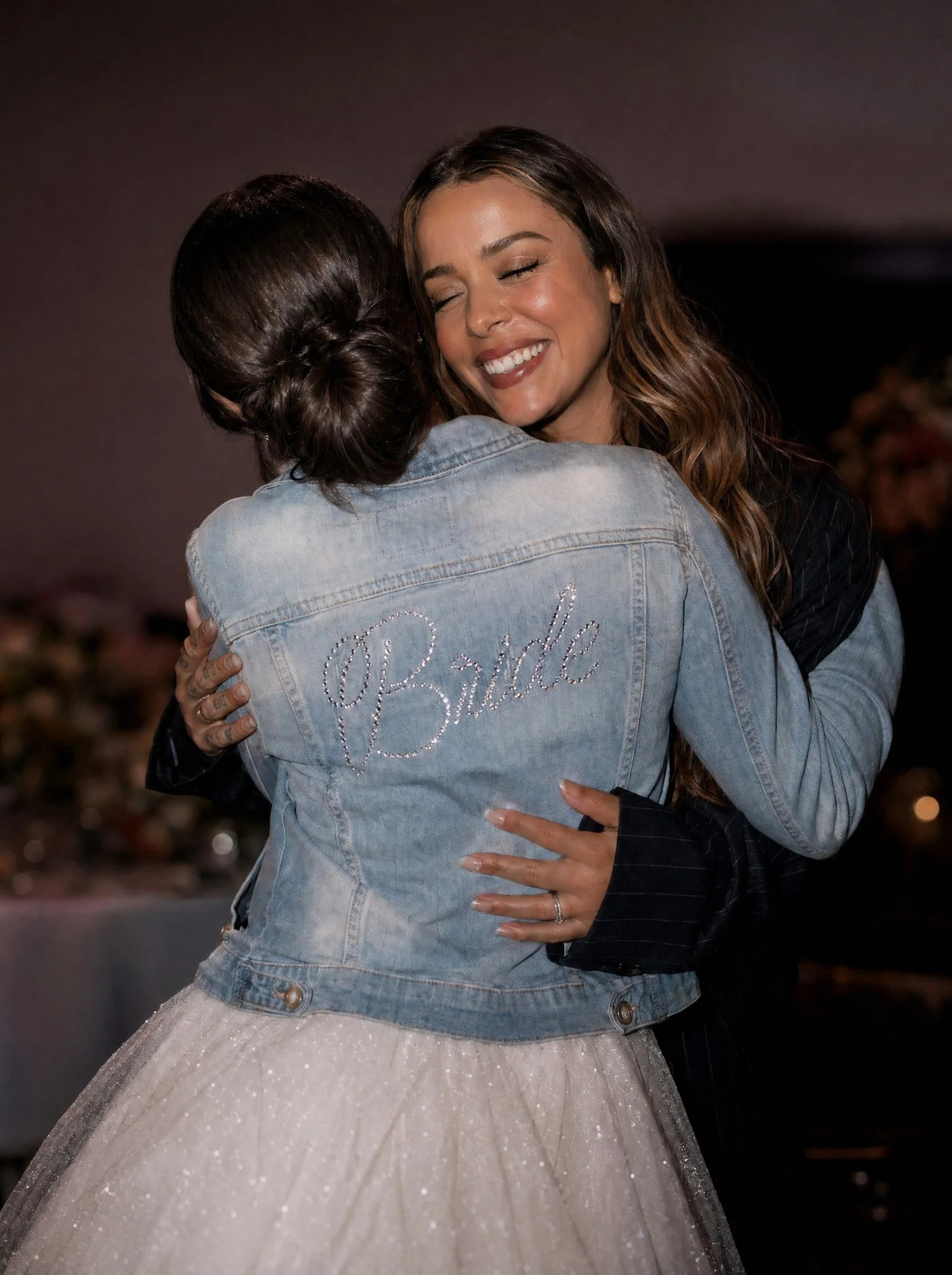 Two women are hugging each other, one in a light-colored wedding dress and the other in a denim jacket with the word "Bride" rhinestones on the back.
