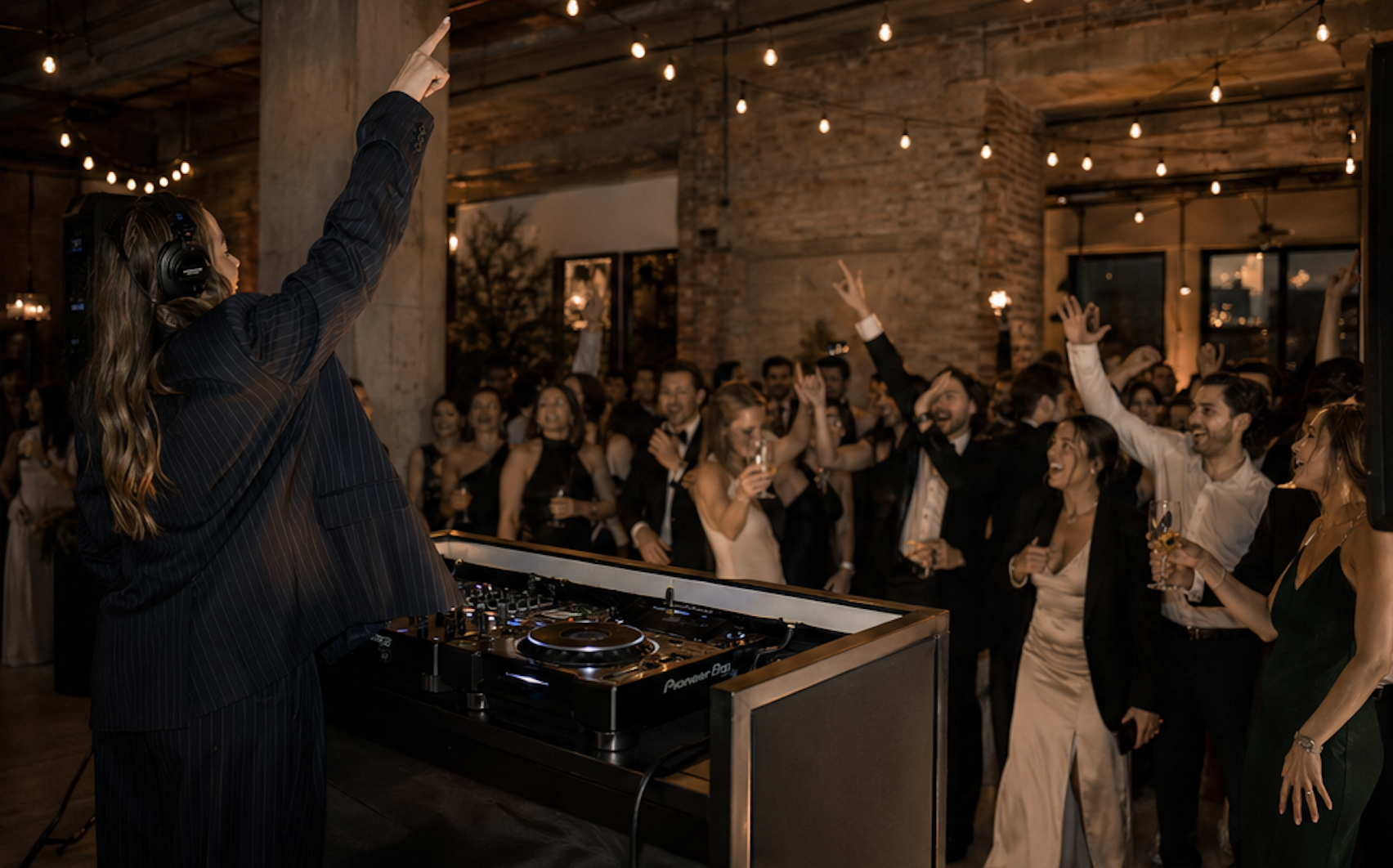 A DJ wearing a black suit and headphones is playing music at a lively party. The crowd, dressed in formal attire, is dancing and having a great time under warm string lights in a rustic indoor venue.