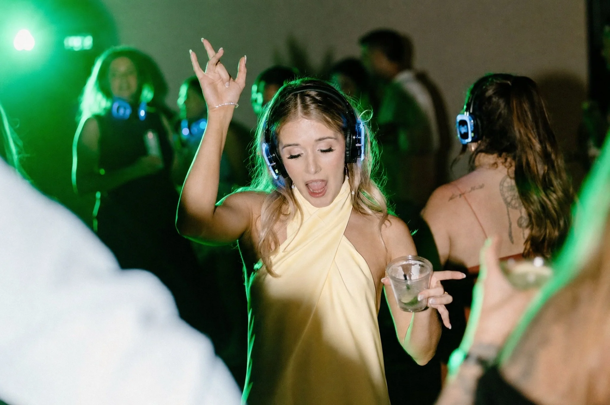 A woman with blonde hair in a yellow dress is dancing with her eyes closed, holding a drink in her left hand, at a party with people wearing headphones and colorful lighting.