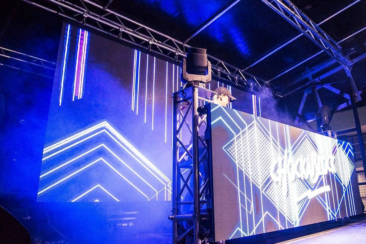 Stage with large digital LED screens displaying geometric blue and white line patterns, supported by a metal truss structure with stage lighting and fog effects.