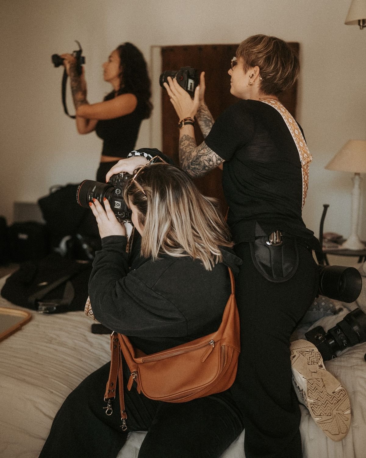 Three women with cameras, two standing and one sitting, taking photographs inside a room with a bed, lamp, and various items on surfaces.