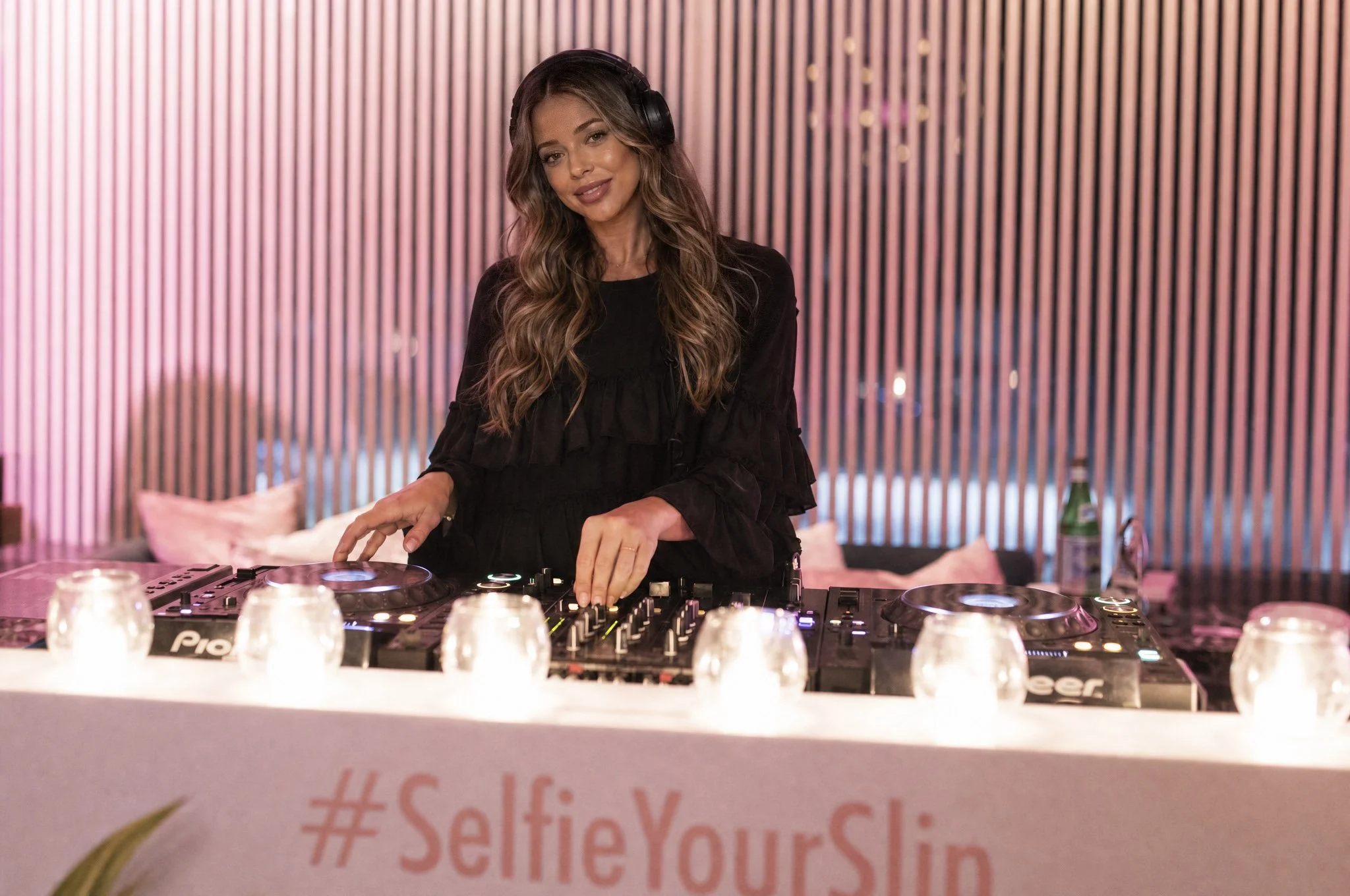 A woman DJ wearing headphones is standing behind a DJ mixer and equipment in a pink-lit room, with candles and a soju bottle on the table in front of her.