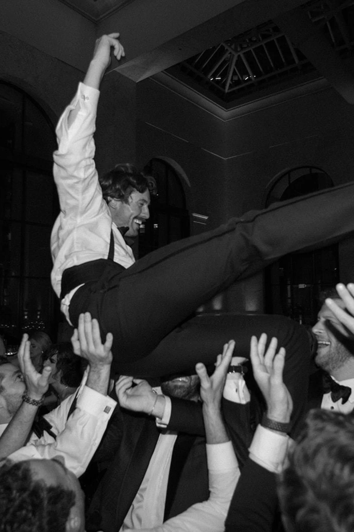A man in formal attire, smiling and with his eyes closed, is being lifted above a crowd of people at a celebration or party. The crowd is supporting him with their hands, and the scene is lively and joyful.